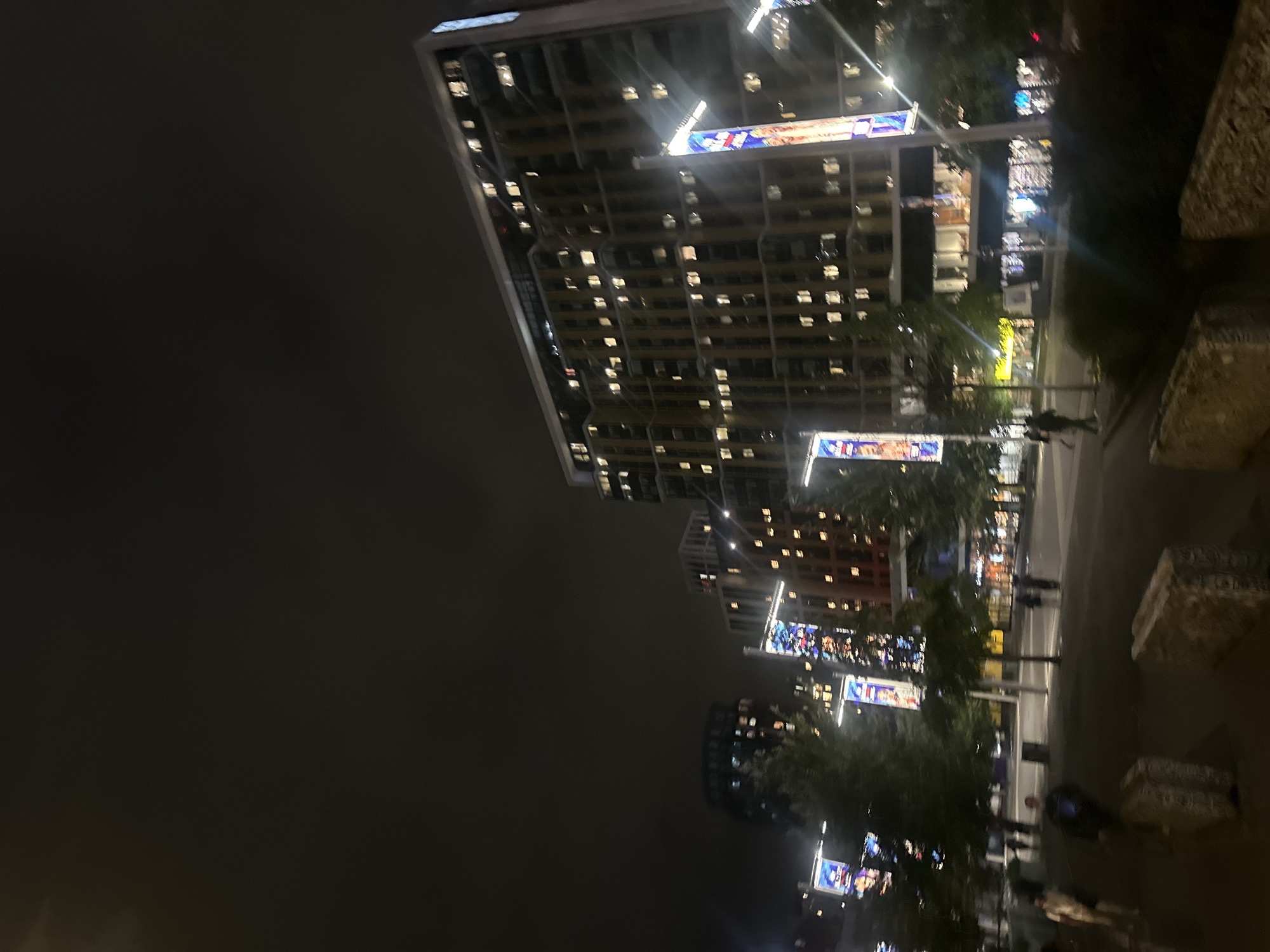 A nighttime, wide-angle, low-light photograph captures a modern urban scene in London, United Kingdom. In the background, a large, multi-story building with numerous illuminated windows dominates the frame, suggesting it's an apartment complex or office building. Smaller buildings, also lit from within, are visible to the left and right of the main structure. Several tall, slender lampposts with colorful banners attached are positioned throughout the scene, casting light on the area. Trees, some appearing as dark silhouettes against the buildings, are scattered across the plaza. In the foreground, several people are walking, with some appearing as blurred figures due to motion or the low light conditions. Concrete barriers are placed on the ground in the very front of the image. The sky is dark and overcast, typical of an evening in London.