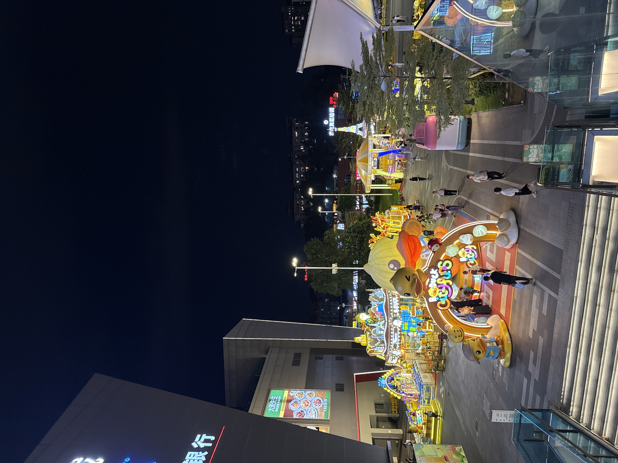 This is an aerial view at night of an outdoor plaza in Tianhe District, China, likely part of a shopping mall or entertainment complex. The plaza features several amusement rides and attractions, illuminated with bright, colorful lights. In the foreground, a wide staircase with integrated lighting descends into a glass-enclosed area. To the left, a large, dark building with prominent neon signage, including Chinese characters that translate to "Bank," is visible.
Dominating the center of the plaza is a large, arched entranceway decorated with cartoon ducks and the text "Big Duck City Fun." Small statues of cartoon ducks are placed around the entrance. Behind this arch, a carousel with multiple levels and numerous illuminated figures, including what appear to be cartoon characters and Paris landmarks, is in operation. To the right of the carousel, a pink and white structure resembling a large toy block or vehicle is parked. Further into the background, more amusement rides, including what looks like a mini-Eiffel Tower with surrounding lights and a Ferris wheel, are visible.
Several people are scattered throughout the plaza, walking and interacting. The ground is paved with a pattern of light and dark gray tiles. Trees and streetlights are also present, and in the distance, apartment buildings with illuminated windows suggest a residential area. The overall atmosphere is festive and vibrant, typical of a nighttime entertainment district.