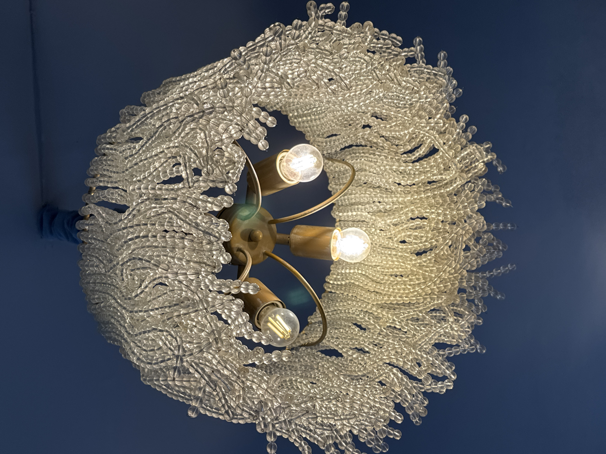 Looking directly up, the photograph showcases a chandelier hanging from a blue ceiling. The fixture features three illuminated light bulbs with coiled filaments, surrounded by a dense arrangement of clear glass beads. The beads form a spherical or circular shape around the light source. The metal components of the chandelier appear to be a brassy color. The photograph provides a close-up, low-angle view of the ceiling fixture. While not explicitly stated in the image, the question implies that it is located in Saint Petersburg, Russia.