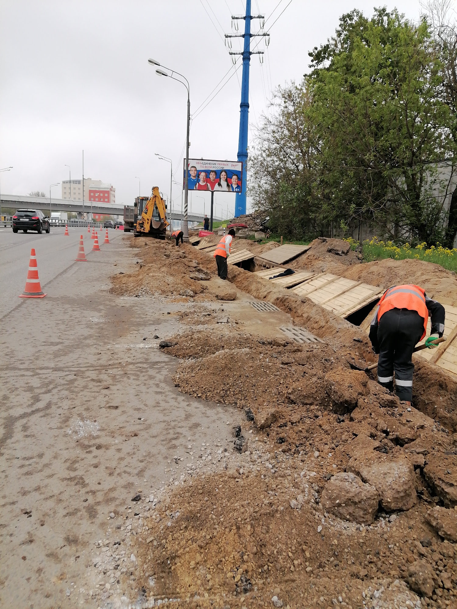 The image depicts a street construction site in Moscow, Russia. Workers wearing orange vests are digging a trench alongside a road marked by orange traffic cones. An excavator is parked nearby. The trench is partially covered with wooden planks. In the background, there's a highway overpass with moving vehicles, a building, a billboard showing faces of people, and a blue electrical support tower. The weather appears overcast.
