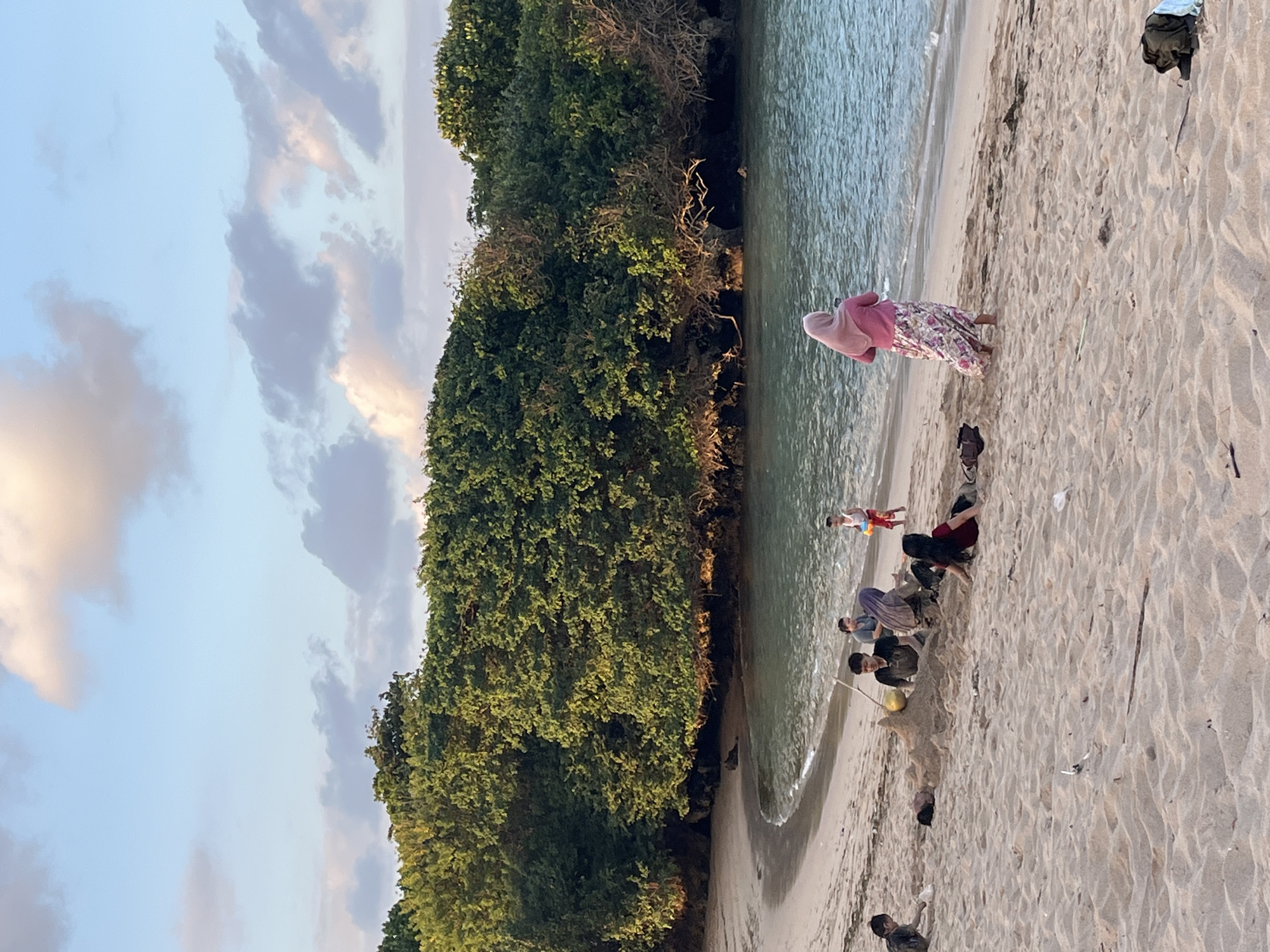 The image depicts a scenic beach with lush green trees lining the background. The foreground is dominated by a sandy beach where several individuals are engaged in various activities. In the center, a group of young people are building a sandcastle, with one individual using a stick to add details. To the right of the sandcastle builders, a young boy in a red and white outfit stands holding a yellow toy bucket and shovel. Further to the right, a person wearing a pink hijab and a floral skirt stands facing the ocean, appearing to be holding a phone. On the left side of the image, a young child is playing in the sand. The ocean is visible in the mid-ground, with gentle waves lapping at the shore. The sky above is a soft blue with scattered clouds, suggesting it might be late afternoon or early evening. The overall scene conveys a relaxed atmosphere of a family or group enjoying a day at the beach. This is likely Kuta Selatan, Indonesia, based on the presence of similar beaches and vegetation.