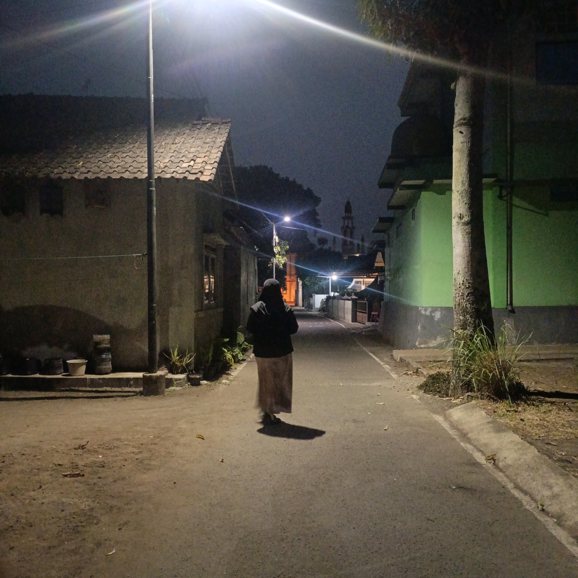 The image depicts a dimly lit, narrow street in Salatiga, Indonesia, at night. A single person, viewed from behind, walks away from the camera down the asphalt road. The person is wearing a dark hijab and a long, light-colored skirt. A tall tree with a rough trunk stands prominently to the right of the person, casting a shadow. On the left side of the street, a building with a tiled roof and rough-textured walls is visible. The building has a window with light emanating from within. The right side of the street is lined with a building painted a vibrant green, with a gray concrete base. Streetlights illuminate the scene, creating lens flares and highlighting the road and the surroundings. The ground on the sides of the road is dirt and sparse vegetation. In the background, a mosque minaret is visible against the dark night sky. The overall atmosphere is quiet and solitary.