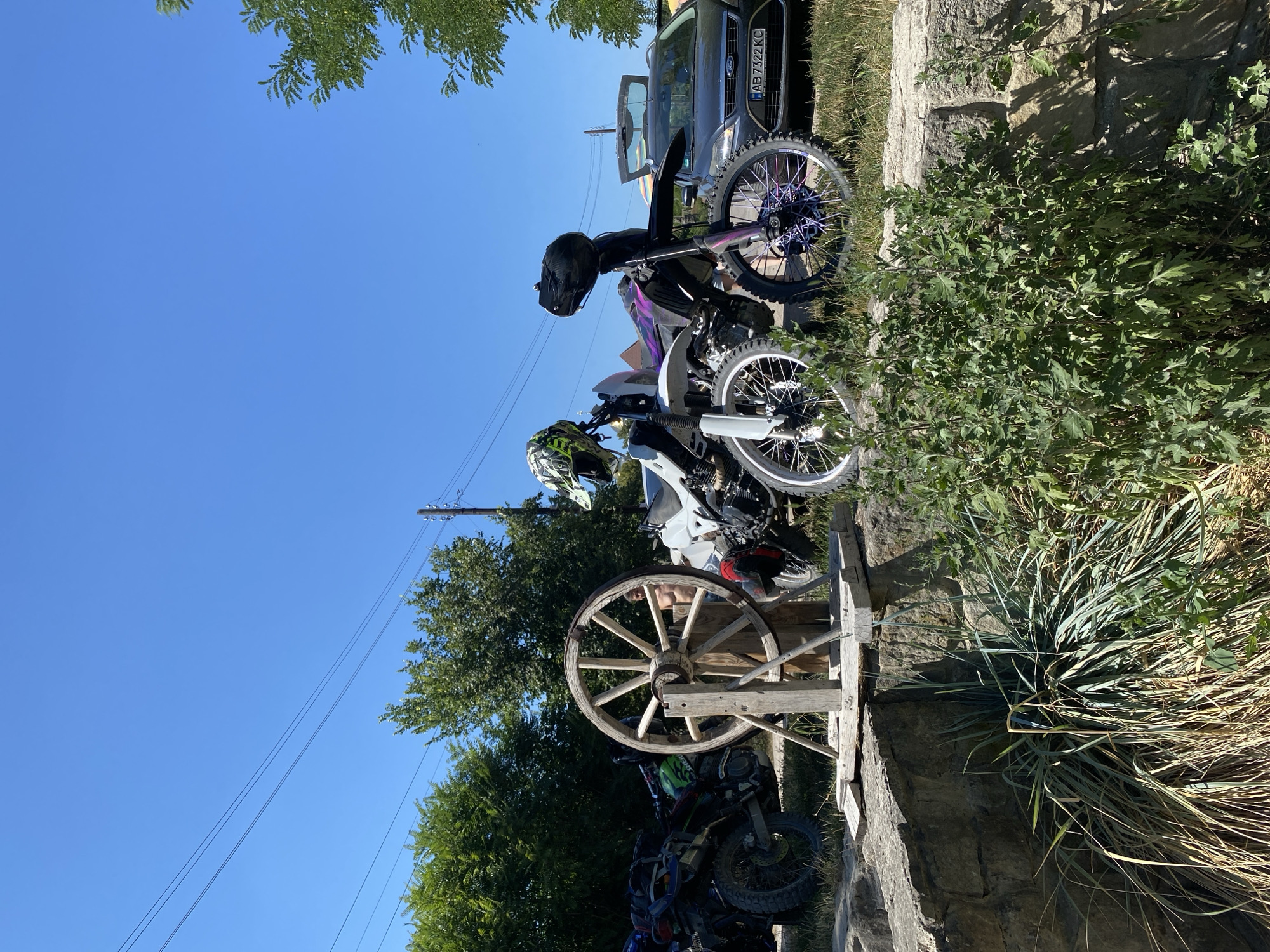 In the city of Busha, Ukraine, a bright, clear blue sky dominates the upper portion of the frame, with thin power lines crisscrossing the expanse. Below the sky, lush green trees form a dense backdrop. In the foreground, a rustic scene unfolds with several motorcycles and a dark-colored car parked on a grassy area.
Prominently featured is a large, weathered wooden wagon wheel mounted on a simple wooden stand. To its left, a dark-colored motorcycle with saddlebags is parked, and another motorcycle, green and black, is partially visible behind it. To the right of the wagon wheel, two dirt bikes are positioned. One is white with purple accents on its spokes, and the other is black with purple accents. Both dirt bikes have helmets placed on their handlebars. The black car, a Ford Mondeo, has its trunk open, revealing the interior. The license plate of the car reads "AB 7322 KC." The ground is covered with grass and a low stone wall runs along the edge of the foreground, with a cluster of dry grasses and green bushes growing near it. A shirtless man, his back to the camera, is partially visible standing behind the wagon wheel, looking towards the motorcycles. The overall impression is a gathering of motorcycles and vehicles on a sunny day, with elements of rustic decoration and natural landscape.