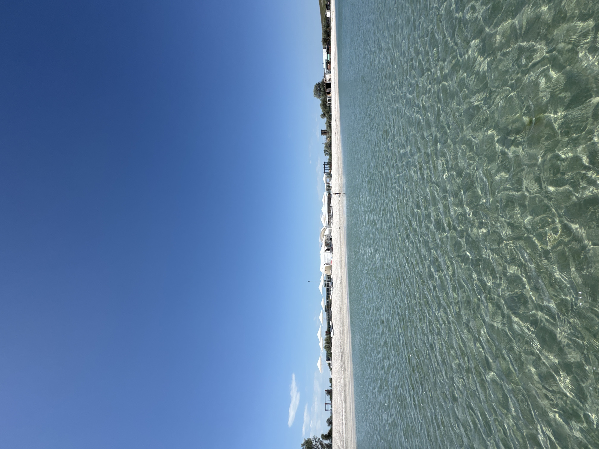 The image depicts a beach scene in Мысовское сельское поселение, Ukraine. The photograph is oriented with the horizon running vertically. The left half of the image is a clear, bright blue sky. The right half shows a white sand beach and clear, shallow water with visible ripples and the seabed. Along the beach, there are several white beach umbrellas. In the distance, there are buildings and trees. A single person stands on the beach in the distance.
