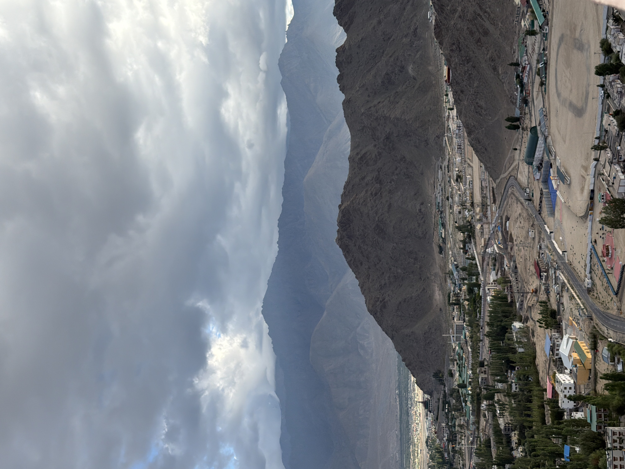 This is a wide, high-angle, landscape photograph of Leh, India. The image is dominated by a dramatic cloudy sky, with patches of sunlight breaking through. In the middle ground, a range of rugged, brown mountains rises sharply from the valley floor. The town of Leh is visible nestled at the foot of these mountains, characterized by a dense collection of buildings with a mix of white, yellow, and red roofs, interspersed with green trees. A winding road is visible, leading through the urban area. In the foreground, the lower slopes of a mountain are covered with sparse vegetation. The overall impression is of a high-altitude, arid environment with a developing urban center.