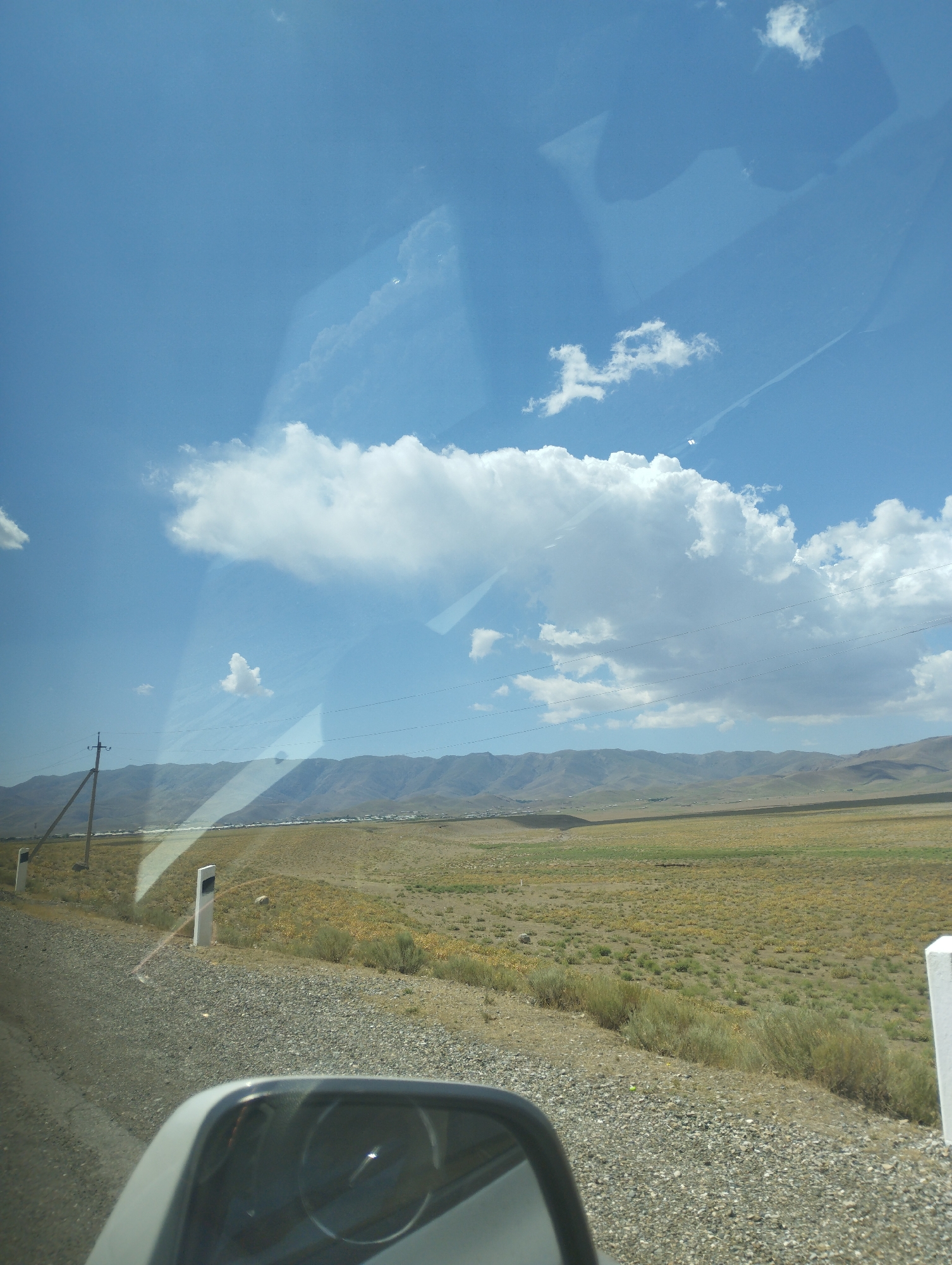 The image captures a view from inside a vehicle looking out towards a vast, arid landscape under a bright blue sky with scattered clouds. In the foreground, a car's side mirror reflects a portion of the interior and the road ahead. The road itself appears to be gravel, with the shoulder marked by white posts. Beyond the road, the terrain transitions into dry, yellow-green vegetation, characteristic of a steppe or semi-desert environment. In the middle ground, a line of trees and buildings suggests a settlement or village, stretching towards a range of low, rolling mountains in the background. A solitary utility pole with wires is visible on the left side of the frame, indicating infrastructure in the area. The scene is consistent with the rural outskirts of Forish QFY, Uzbekistan, known for its expansive natural landscapes and agricultural settlements.