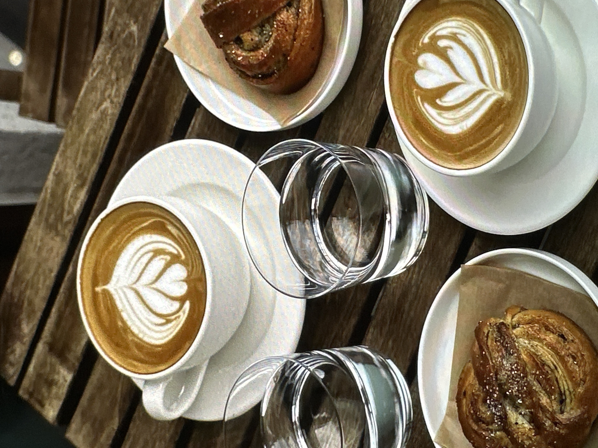The image is an overhead shot of a wooden table with several items on it. There are two cups of coffee with latte art on white saucers. Additionally, there are two plates, each containing a pastry. Between the coffee and pastries, two empty drinking glasses are placed. The location cannot be verified as Yerevan, Armenia without additional information.