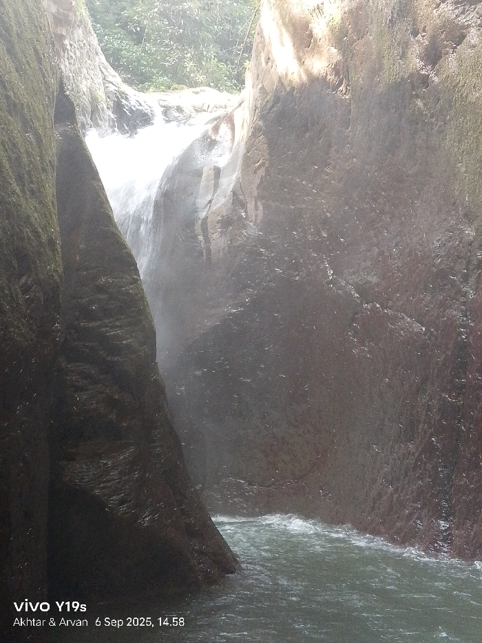 The image displays a natural waterfall within a narrow, rocky canyon, specifically located in Sukadana, Indonesia. A significant volume of white, turbulent water descends from an upper opening, generating visible mist and spray, into a flowing, greenish-blue river at the canyon's base. The canyon walls are steep and prominent; the left wall is dark, textured, and covered with moss, extending into the lower left foreground. The right wall, also rocky and wet, exhibits streaks and lighter brown-grey tones from water runoff. Dense green foliage is visible at the top edge of the canyon, above the waterfall's source. The river in the foreground shows active currents and ripples as it flows away from the waterfall. The scene is naturally lit, with bright highlights on the falling water and upper rock surfaces contrasting with the more shadowed canyon interior. No human figures or man-made objects are present within the natural environment depicted.