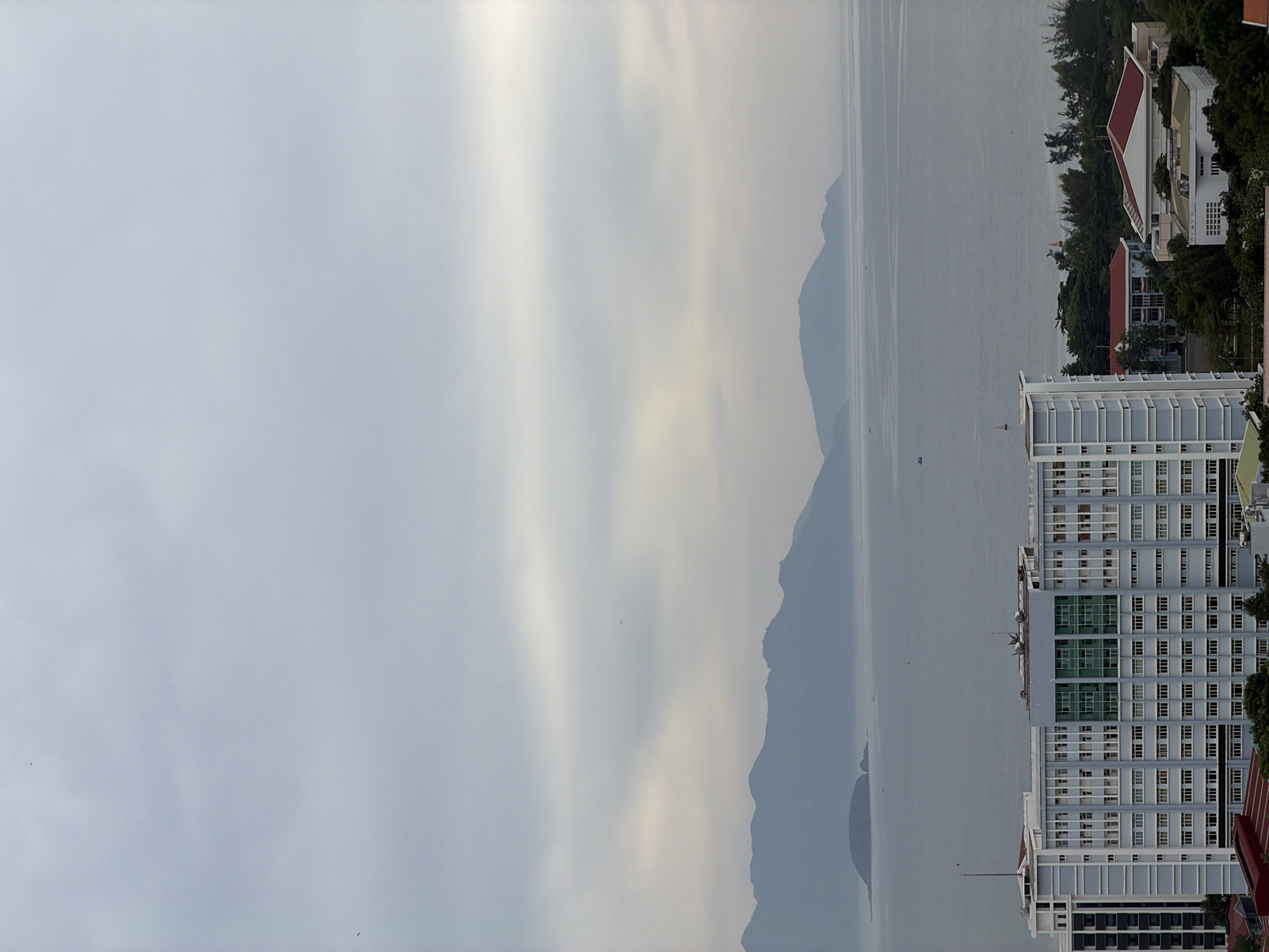 This is a high-angle, full-frame shot of the city of Nha Trang, Vietnam, under an overcast sky. The image captures a vast expanse of calm, gray water, likely the ocean, stretching towards a distant, hazy mountain range. In the foreground, several buildings are visible. The most prominent is a large, light blue and white multi-story building with numerous windows, featuring a section with green-tinted windows. To its left and right, other buildings and lush green trees are present. A few small boats are scattered across the water. The overall scene suggests a peaceful, possibly early morning or late afternoon setting due to the soft, diffused light.