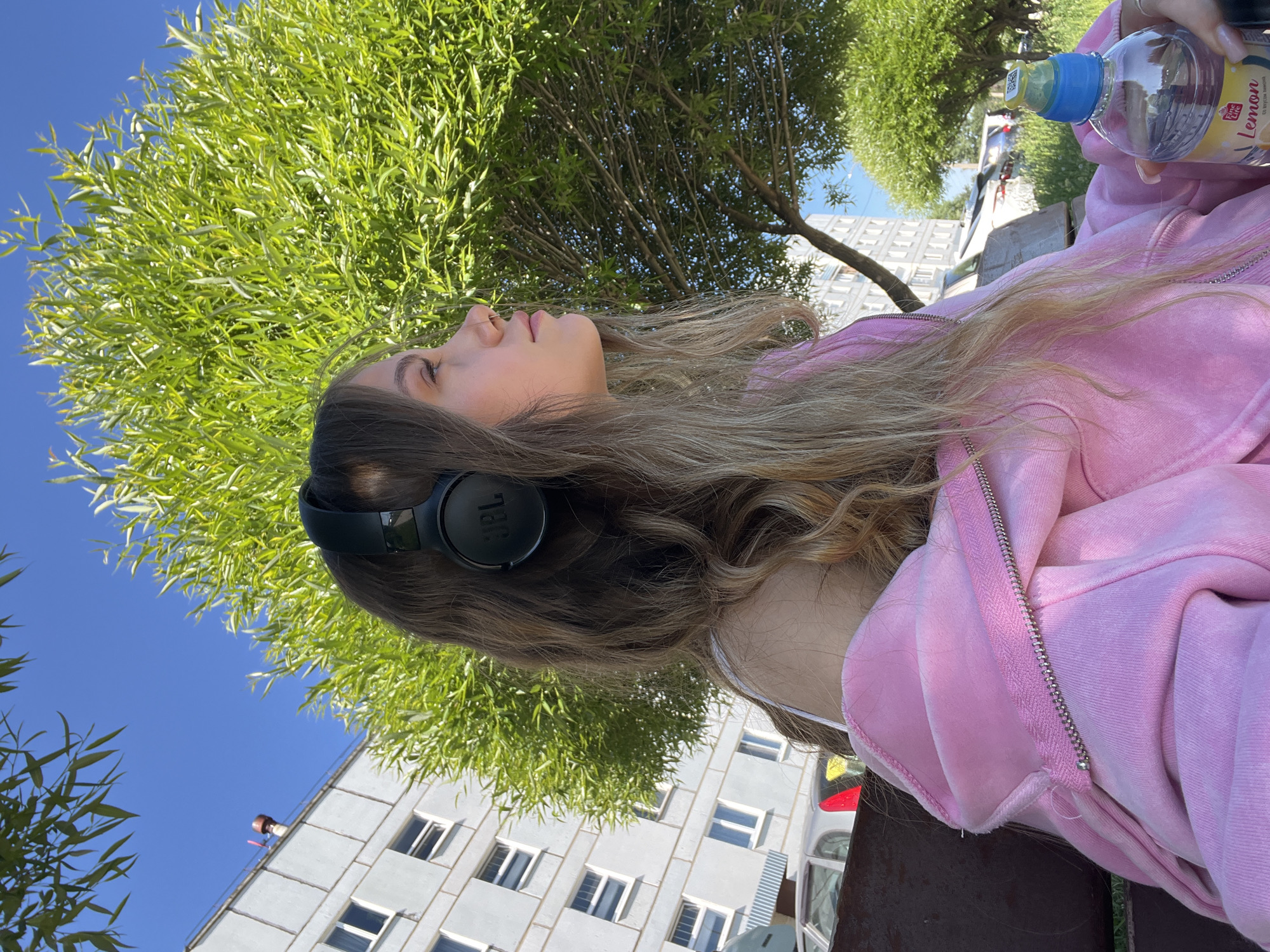 A young woman with long, wavy blonde hair and wearing black headphones sits outdoors on a bench in the city of Chaikovsky, Russia. She is looking to her right, with her profile visible. She is wearing a light pink hooded jacket, partially unzipped. In her right hand, she holds a clear plastic bottle of water with a yellow label that reads "Fine Life Lemon." Behind her, a large green tree with lush foliage dominates the frame. In the background, a multi-story apartment building with white facades and evenly spaced windows is visible, along with parked cars and other greenery. The sky is clear and blue, indicating a sunny day.