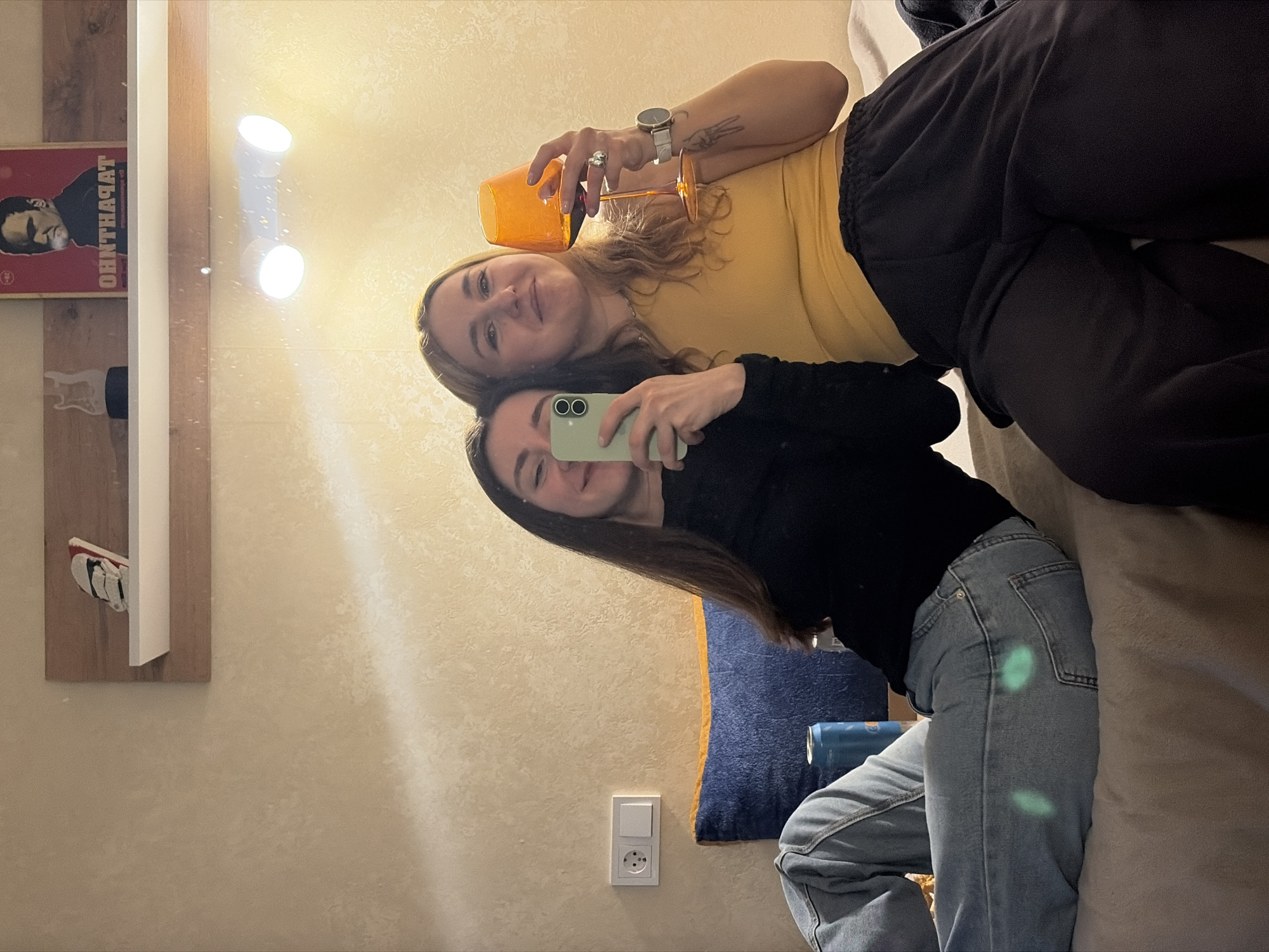 Two young women pose in a mirror selfie. One is wearing a yellow top and dark sweatpants, holding an orange glass. The other is wearing a black top and blue jeans, holding a light green phone. They appear to be in a room with light-colored walls. Visible in the reflection are decorative items on shelves, a blue pillow, an electrical outlet, and a can of drink. The location is identified as Unknown, Unknown.
