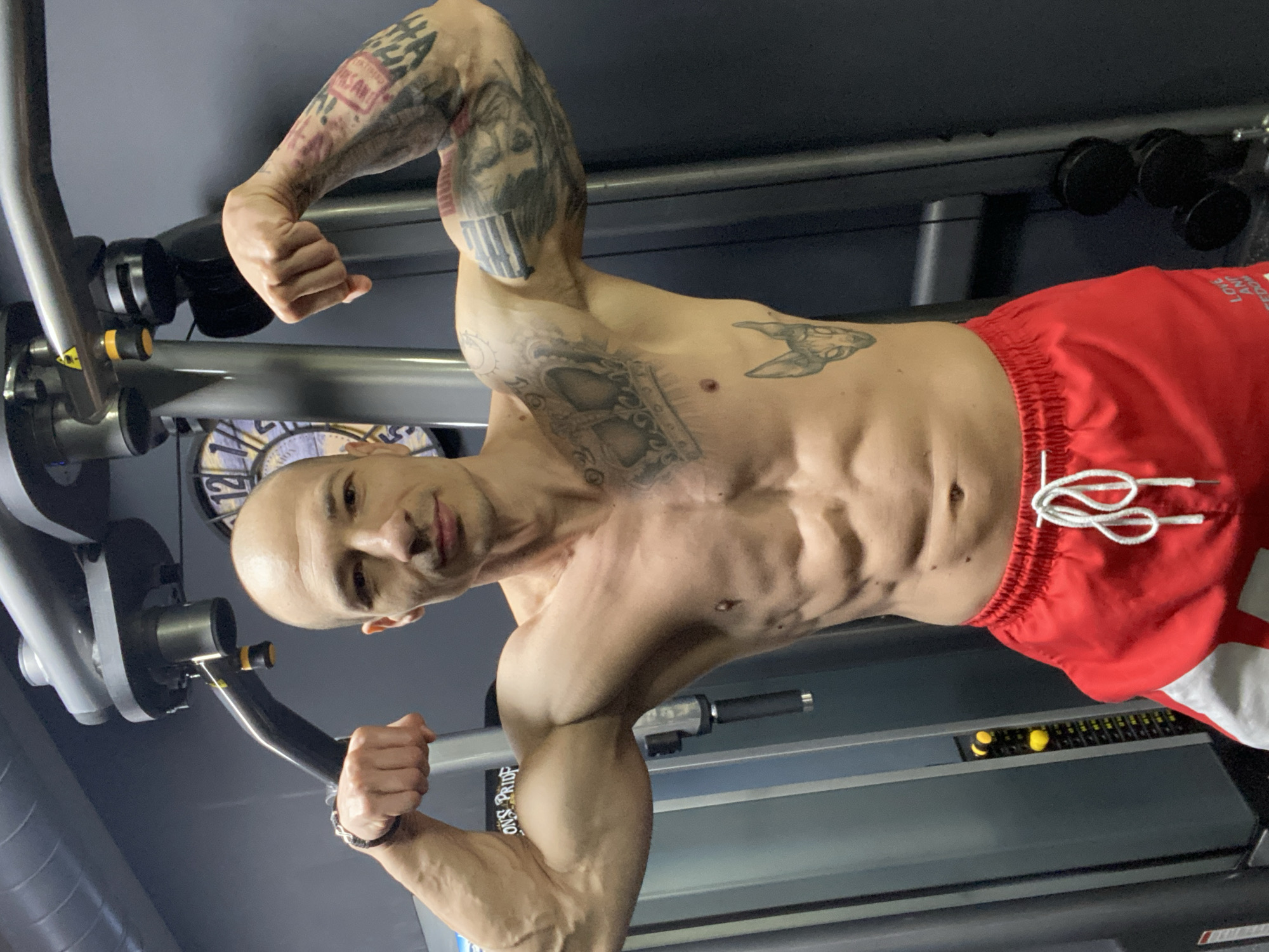 A shirtless man with a shaved head and extensive tattoos is posing in what appears to be a gym. He is wearing red shorts with white drawstrings and a white stripe on the side. He is flexing his arms, revealing well-defined biceps and abdominal muscles. He is holding onto the handles of a weight machine, possibly performing a chest exercise. The background shows gym equipment, including weights and a clock. The exact location within Pleven, Bulgaria cannot be determined from the image.