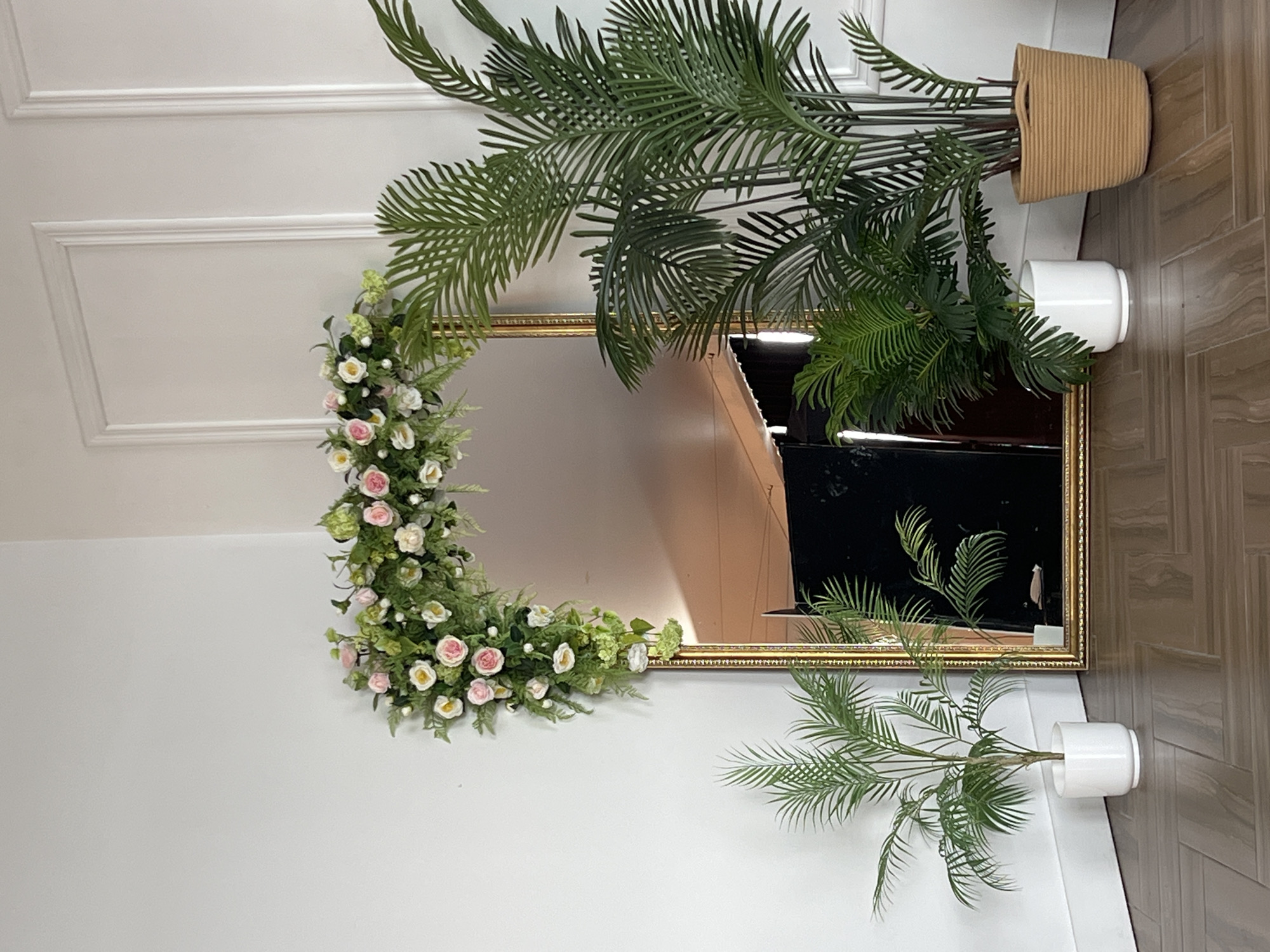 A mirror is decorated with flowers and surrounded by plants in pots. The scene is indoors.