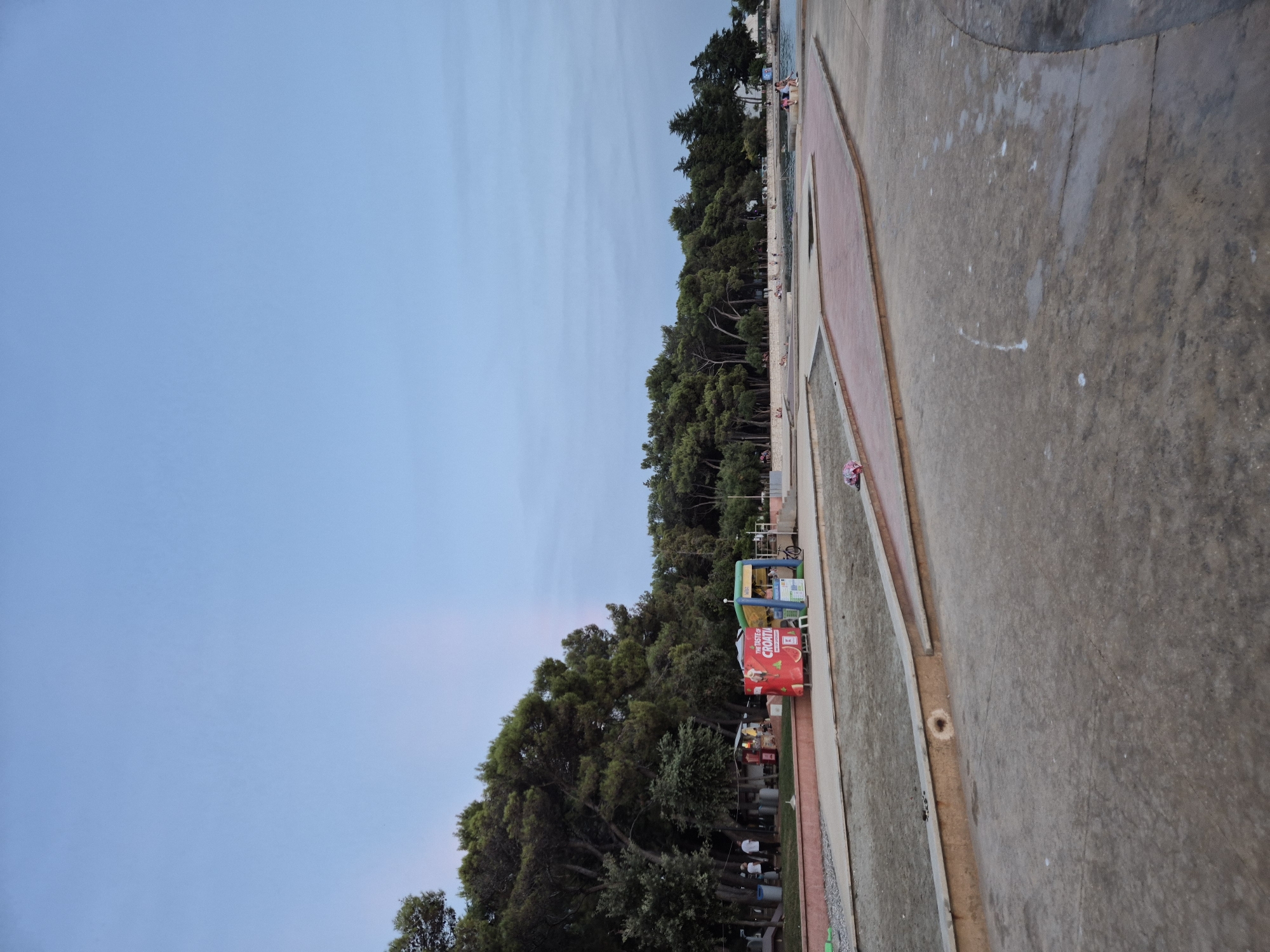 The image depicts an outdoor scene with a clear blue sky. In the foreground, there is a paved area with a textured surface, appearing to be a walkway or plaza. Several concrete or stone-like barriers are visible, creating distinct sections within the paved area. Some of these sections contain sand or dirt.
In the middle ground, there is a line of trees forming a dense, green backdrop along a slightly elevated area. Among the trees, several small stalls or kiosks are visible. One prominent stall has a red exterior with a graphic of a sliced fruit, and text that includes "THE TASTE OF CROATIA". Next to it is a blue and green inflatable structure. A few people are present, some standing near the stalls and others walking.
Further back, there are more trees and an area that appears to be water, possibly a bay or a pool, with a few people sitting or walking near its edge. The overall environment suggests a coastal or recreational area. The lighting indicates it is likely daytime, but possibly late afternoon or early morning due to the soft light. The scene is identifiable as being located in Katoro, Croatia.