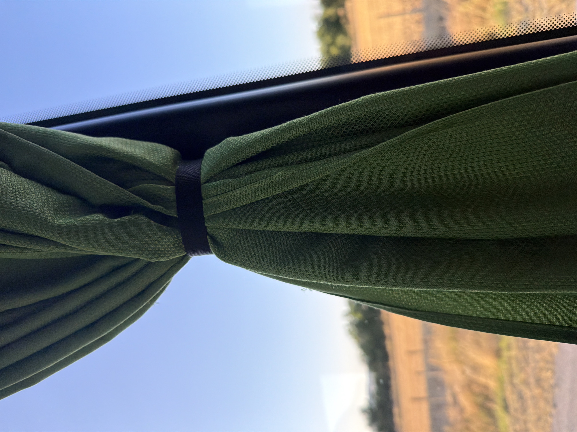 A green curtain is tied back with a black strap, obscuring a portion of a window that looks out onto a rural landscape. The landscape features dry, yellow grass and a line of trees in the distance under a clear blue sky. The image appears to be taken from inside a vehicle, as indicated by the texture of the windowpane visible along the right edge. There are no people or animals in the image. The location in the city of Shtarkovo, Bulgaria, is not discernible from the visual information presented in the image.