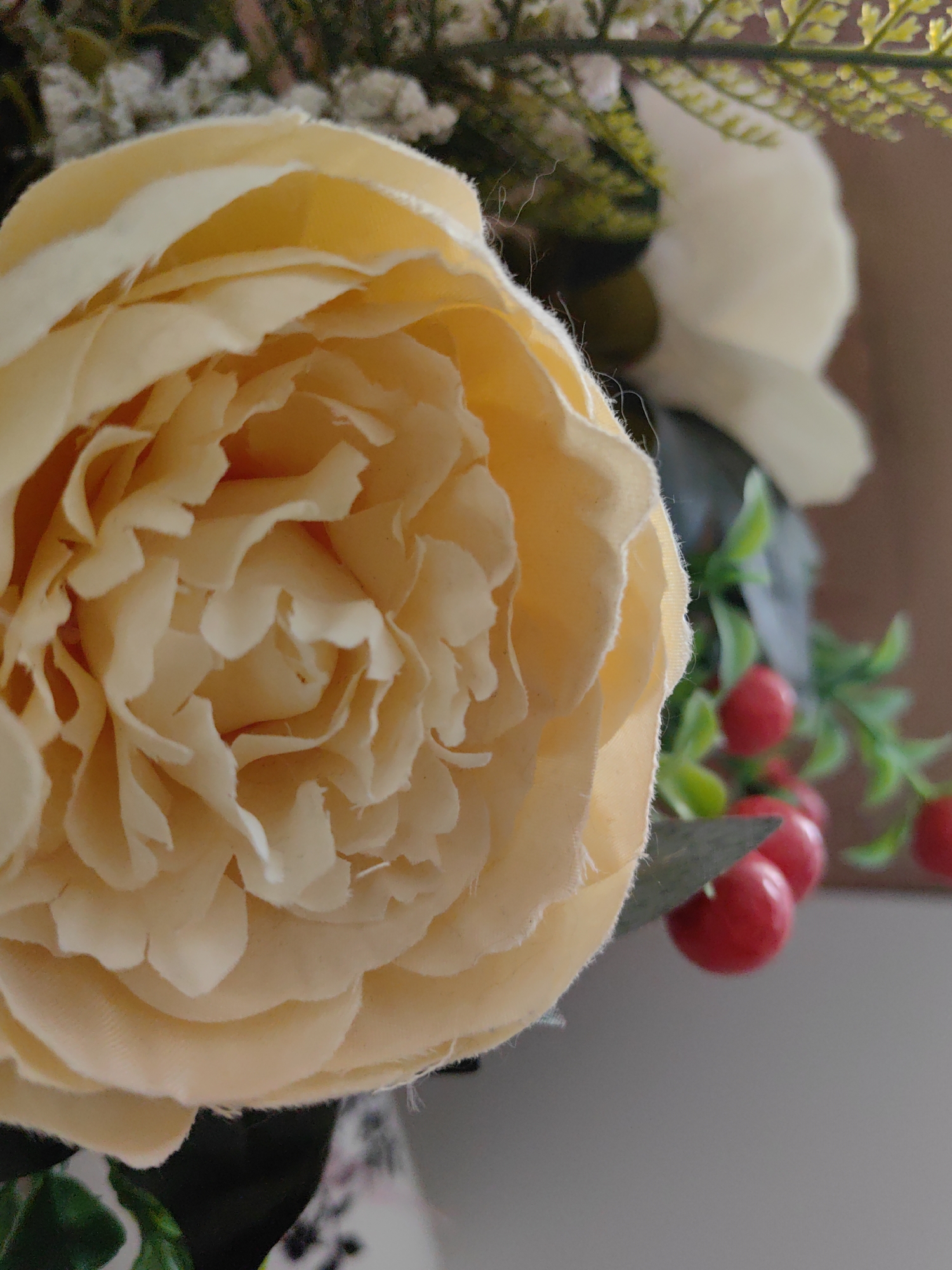 The image displays a close-up of an artificial floral arrangement. A large, light yellow or cream-colored artificial flower, resembling a peony or rose with fabric petals exhibiting slightly frayed edges, occupies the majority of the left and central frame. To the upper right, a portion of another white artificial flower is visible, out of focus. Interspersed throughout the arrangement are green artificial foliage elements and clusters of small, round, bright red artificial berries with a glossy finish, predominantly located on the right side. The background is blurred, featuring indistinct warm brown tones in the upper right and lighter gray tones in the lower right. No individuals or specific identifying location details are present.