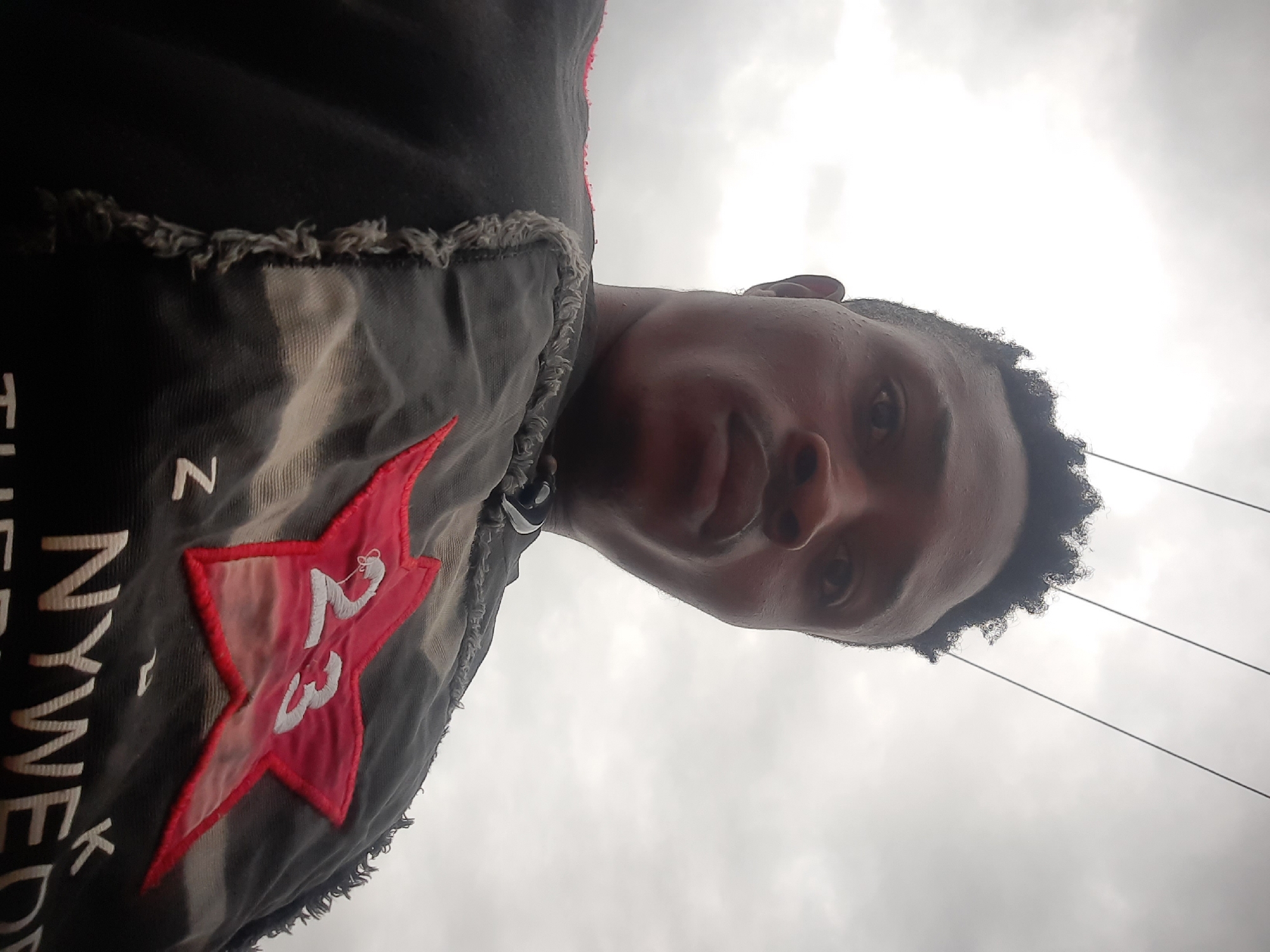 A low-angle self-portrait of a dark-skinned person. The subject's face is centered, with their eyes looking down at the camera. They have short, dark, curly hair. They are wearing a dark-colored garment with a red star emblem featuring the number "23" and the partially visible text "NYWEDK" on the left side of the frame. The background shows an overcast sky and several utility lines. This image was taken in Jos, Nigeria.