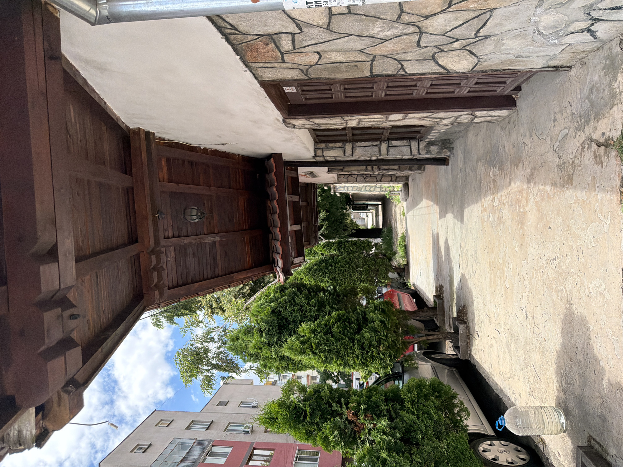The image depicts an outdoor scene in Pazardzhik, Bulgaria, likely on a street with buildings and parked cars. A covered walkway with a wooden roof and exposed beams runs along the right side of the frame, adjacent to a building with a stone facade and a brown wooden door and windows. Several coniferous trees are planted in a row along the left side of the walkway, with cars parked behind them. A large plastic water bottle is visible on the paved ground in the foreground, to the left of the walkway. In the background, a multi-story apartment building is visible, along with more trees and a glimpse of the sky with clouds. The overall impression is of a residential or commercial street scene.