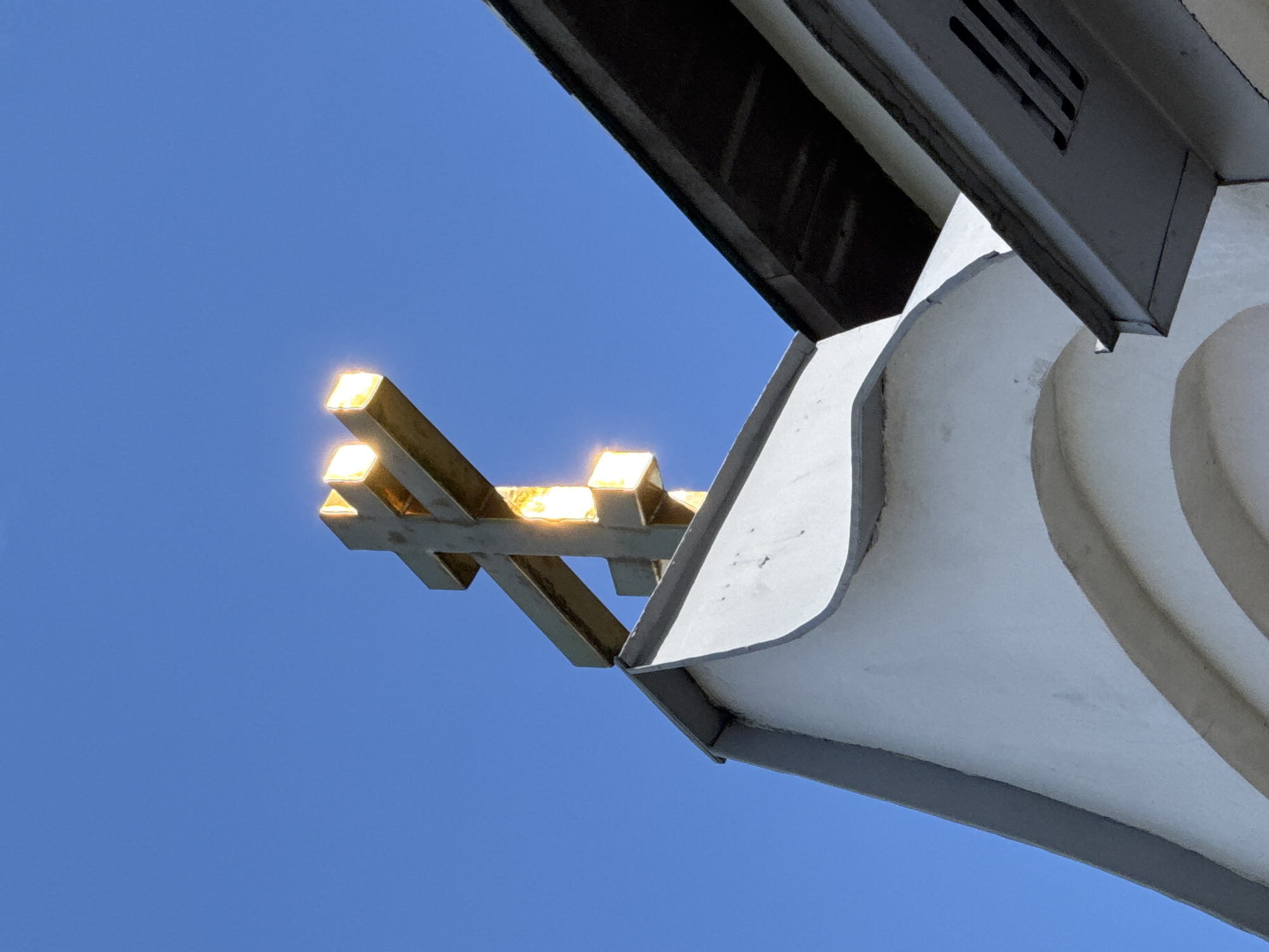 The image shows a close-up, low-angle view of a gold-colored, triple-barred cross atop a white building. The cross is brightly lit by the sun, causing some parts to appear overexposed. The background is a clear, vibrant blue sky. Part of a dark-colored roof structure is visible to the right of the cross. The building beneath the cross has a white, slightly textured facade with a curved architectural element. This image was taken at the Alexander Nevsky Cathedral in Saint Petersburg, Russia.