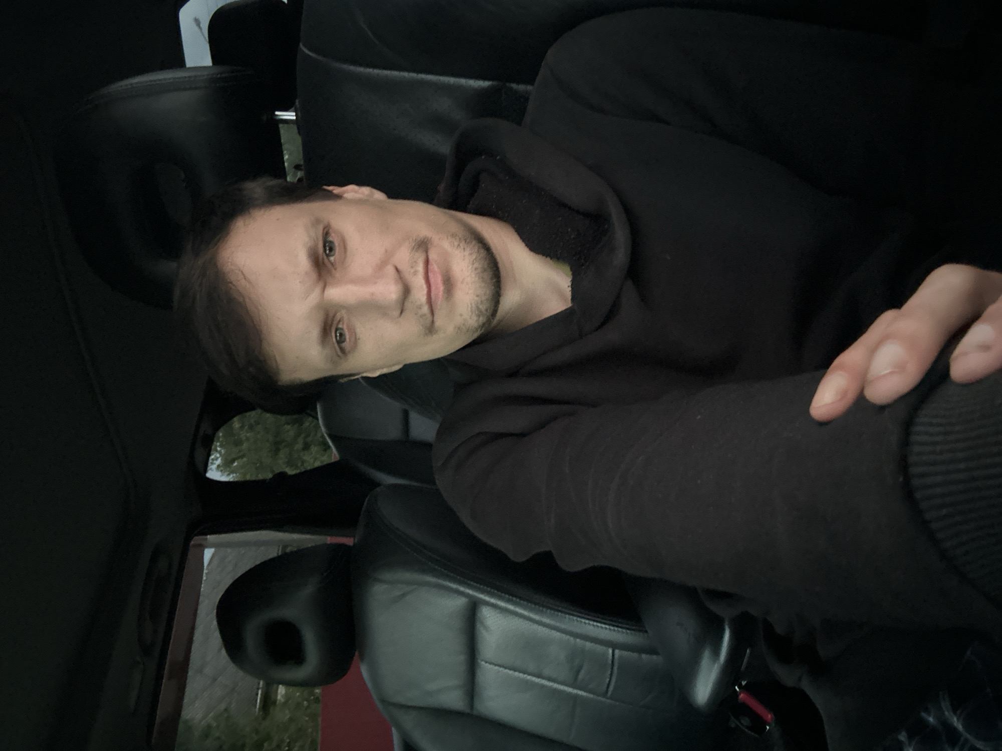 A medium shot is captured from inside a car, looking towards a man sitting in the driver's seat. He has light skin and dark hair, a light beard, and is wearing a black hoodie. The interior of the car is visible, with black leather seats and headrests. The background outside the car is blurry but shows trees and a building. The image was taken in Makiv, Ukraine.
