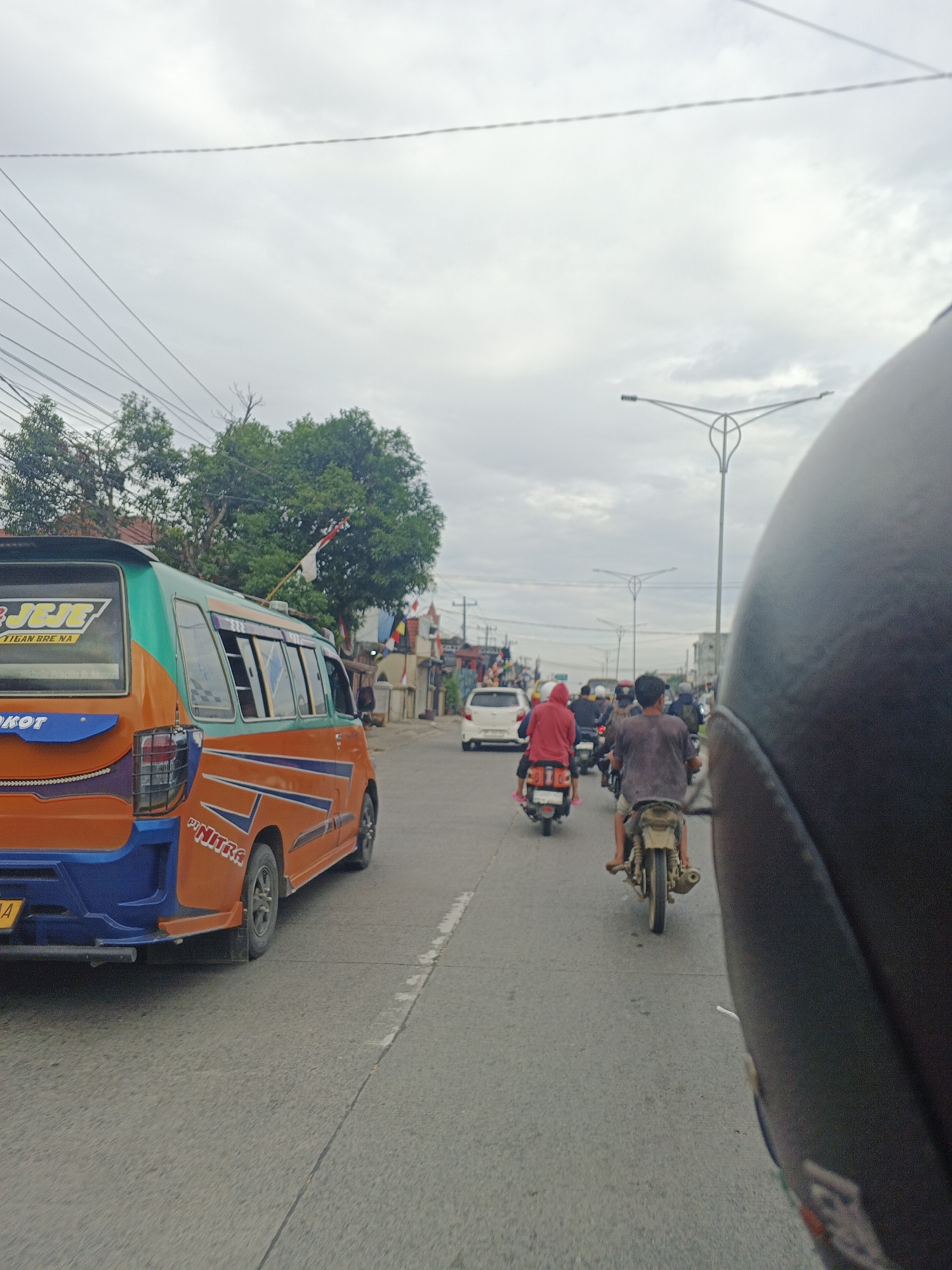 The image depicts a street scene in the city of Medan, Indonesia. The perspective is from the back of a motorcycle, with a dark helmet dominating the right side of the frame. In the foreground, a brightly colored orange and blue van with the word "JEJE" visible on its side is driving in the left lane. The van has various decals and is adorned with a small Indonesian flag on its roof.
The street is paved and marked with white lines. Multiple other motorcycles are also present on the road, forming a traffic flow. One motorcycle in the middle distance carries a passenger wearing a red headscarf. Another motorcycle further ahead has a rider wearing a dark t-shirt. In the background, there are buildings and trees, indicating an urban environment. The sky is overcast with clouds. The overall impression is of everyday traffic in a city.