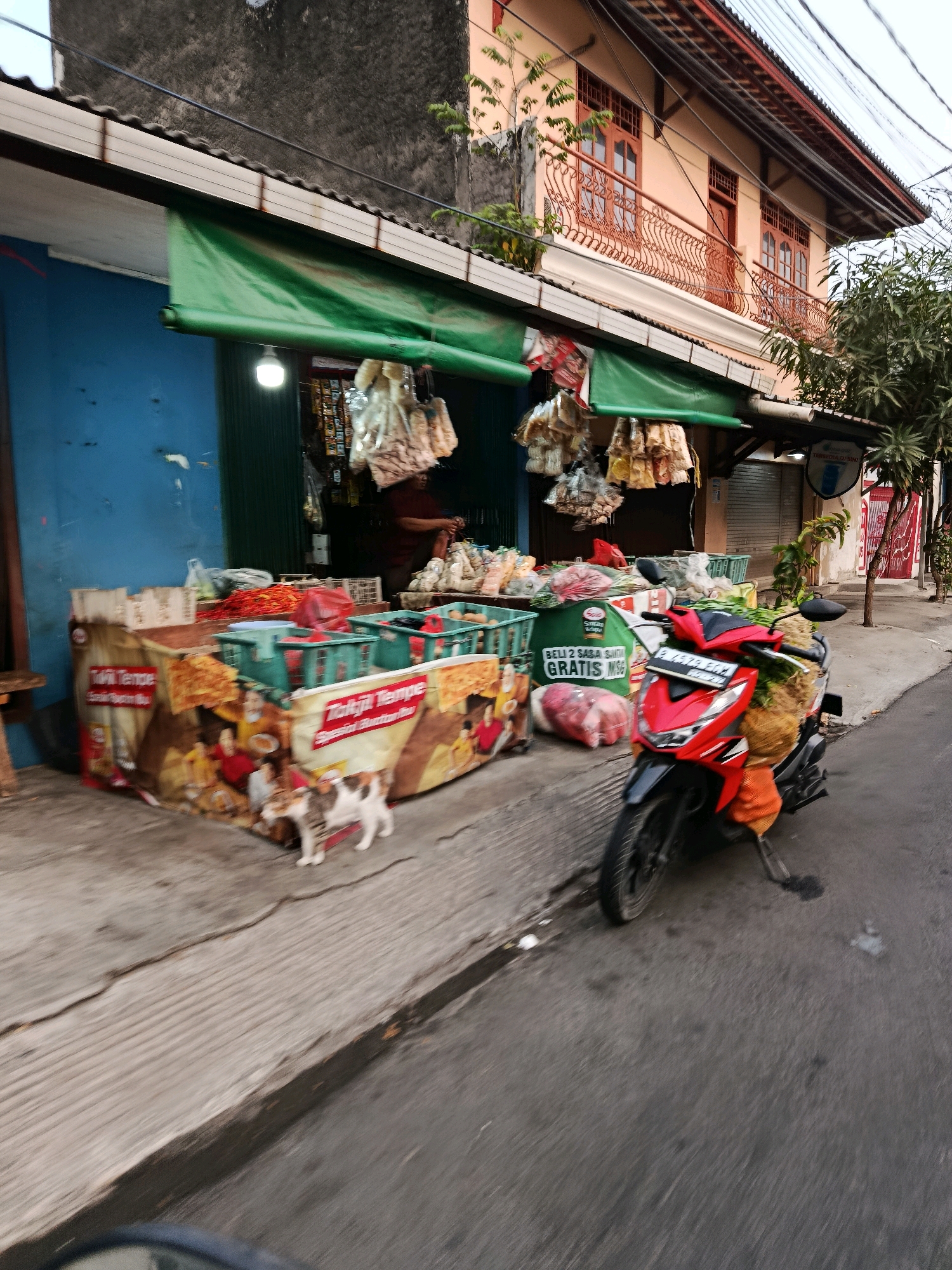 The image depicts a street scene in Bekasi, Indonesia, with a focus on a small roadside market stall. In the foreground, a red scooter is parked on the asphalt road, laden with fresh produce, including large jackfruits and leafy greens. The scooter is positioned to the right of a concrete sidewalk. The market stall itself is set up along the sidewalk and is partially covered by a green awning. Various goods are displayed, including what appear to be dried goods hanging from the awning, baskets of produce, and packaged food items. A prominent display features images of food and text in Indonesian, possibly advertising a product. To the left of the stall, a small, tricolored cat stands on the sidewalk. Behind the stall, the facade of a building is visible, with a blue wall on the left and a peach-colored upper story with ornate balconies on the right. A person, likely a vendor, is partially visible inside the stall, engaged in activity. The environment suggests a typical urban street setting in Indonesia, characterized by the architecture and the presence of street vendors and motorbikes.