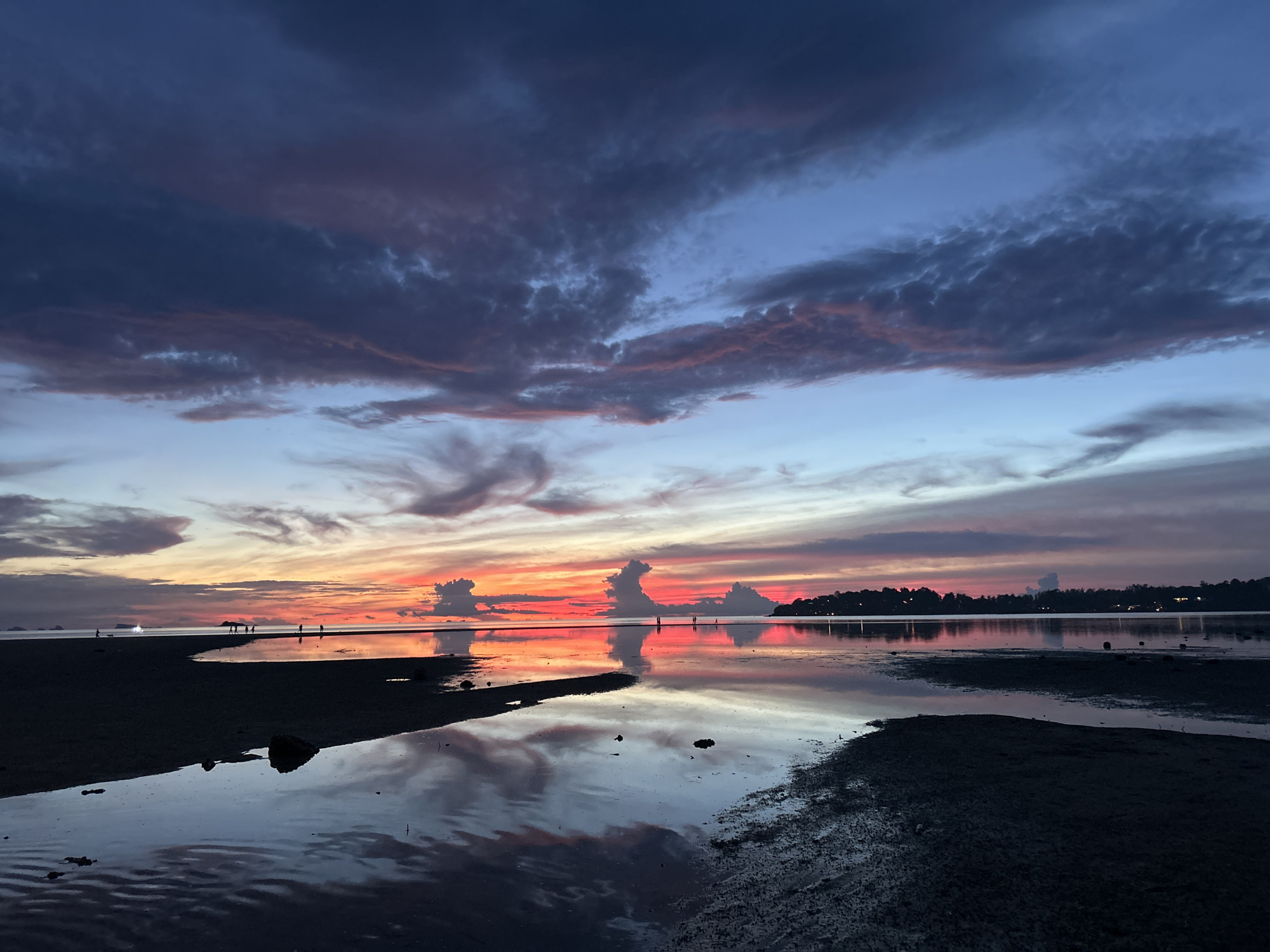 This is a low-angle, wide shot of a beach at sunset in Ko Pha Ngan, Thailand. The sky is a dramatic mix of deep blues, purples, and vibrant oranges and reds, with clouds catching the last light of the sun. The water's surface is calm, acting as a mirror, reflecting the colorful sky and some of the clouds. In the foreground, there is dark sand and shallow pools of water. Silhouetted figures of people can be seen walking along the distant shoreline, and faint lights suggest structures or boats further out. The overall scene is serene and captures the transition from day to night over a coastal landscape.