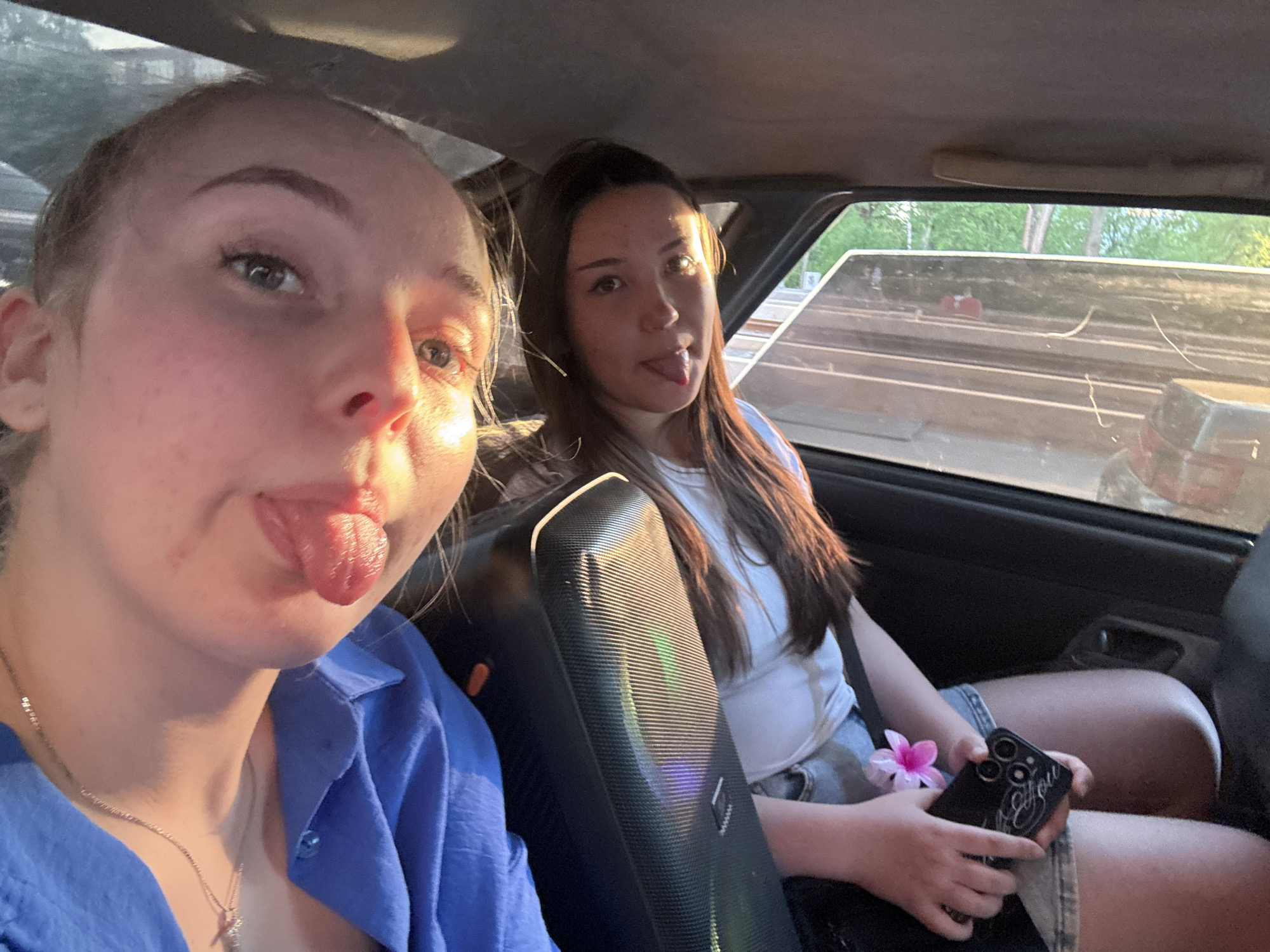 Two young women are in the back seat of a car. The woman in the foreground, wearing a blue shirt, sticks her tongue out. The woman in the background, wearing a white shirt and jean shorts, also sticks her tongue out and holds a phone with a pink flower accessory. A black speaker is visible between them. Outside the car window, there is greenery and a flatbed trailer is visible. The location is likely in Perm, Russia.