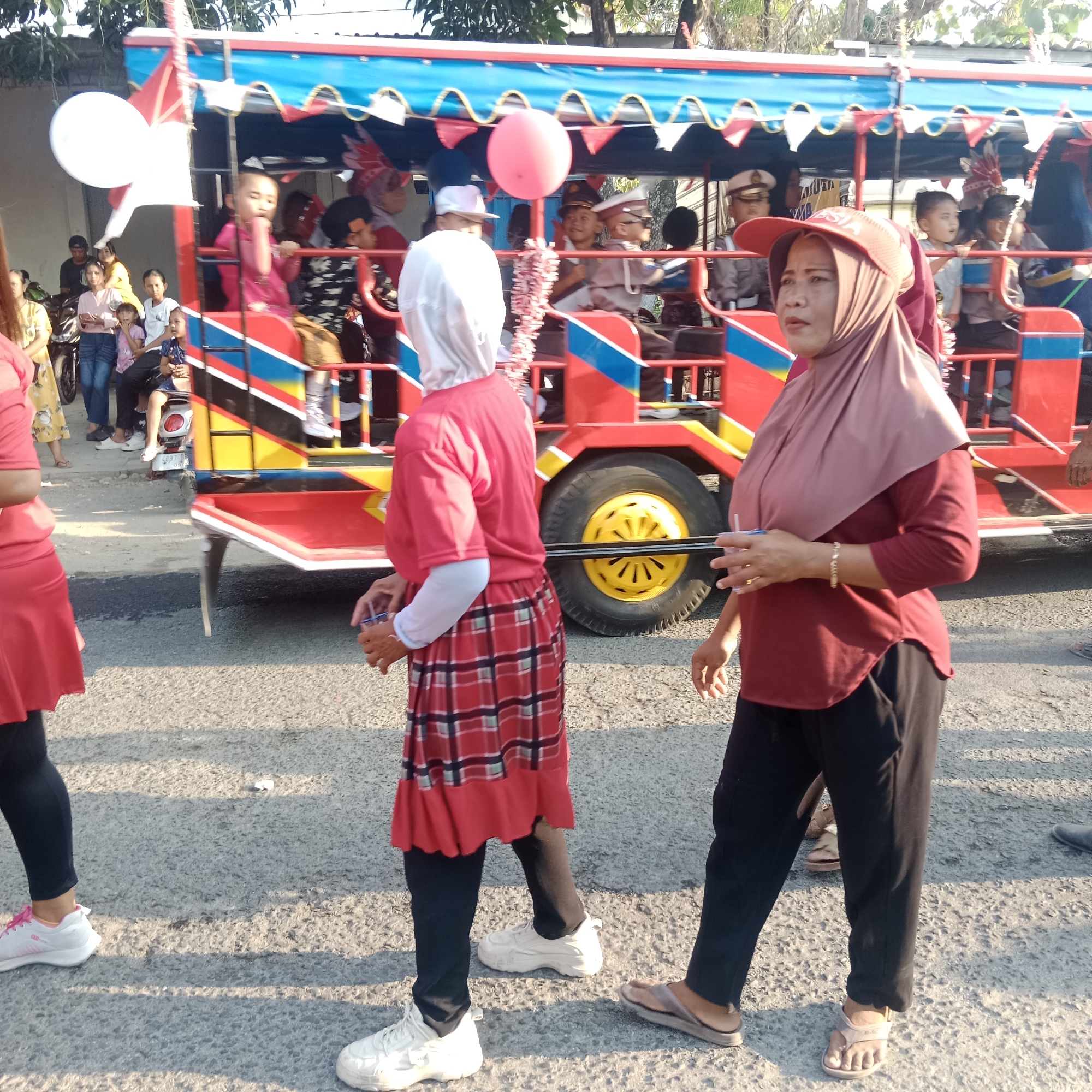 In the city of Tegalrejo, Indonesia, a vibrant parade is underway. A brightly colored open-air vehicle, adorned with red, blue, yellow, and white stripes, serves as the centerpiece. The vehicle is filled with people, including children and adults, some dressed in uniform.
In the foreground, two women are walking alongside the vehicle. The woman on the left wears a red hijab and a red and white plaid skirt over black leggings. She is carrying a clear plastic cup with a straw. The woman on the right wears a maroon hijab and a maroon shirt, with black pants. She has a gold bracelet on her wrist and is also holding a clear plastic cup with a straw.
The street is paved with asphalt, and the warm sunlight casts long shadows. In the background, more people are visible, some standing and watching the parade. Motorcycles are parked on the side of the street. The overall atmosphere suggests a festive community event.