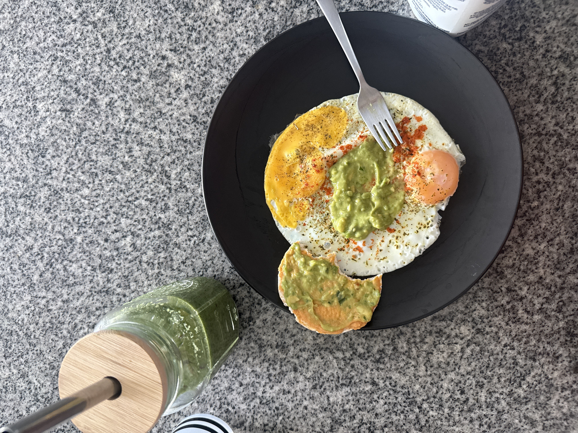 This top-down shot showcases a healthy breakfast or brunch arranged on a speckled gray and white granite countertop. A black, round plate dominates the center, holding two fried eggs. One egg has a fully cooked, bright yellow yolk, while the other presents a softer, orange-yellow yolk. Both are seasoned with dark specks, likely pepper or herbs, and reddish flakes, possibly paprika or chili. A dollop of green mashed avocado, or guacamole, is placed atop the eggs, also seasoned. A silver fork rests on the plate, partially over the eggs.
Beside the plate, a round, flatbread or rice cake, with a bite taken out, is visible. It is spread with the same green mashed avocado. To the left, a clear glass jar with a bamboo lid and a metal straw contains a bright green liquid, likely a smoothie or juice. A fragment of a white and black striped object, possibly a coaster, peeks out beneath the glass. In the upper right background, a white container, possibly for a dairy product, shows partially legible Spanish text, including fragments like "ESPERA" and "LEER." The lighting suggests it is daytime, and the scene implies an indoor setting, possibly a kitchen or dining area.