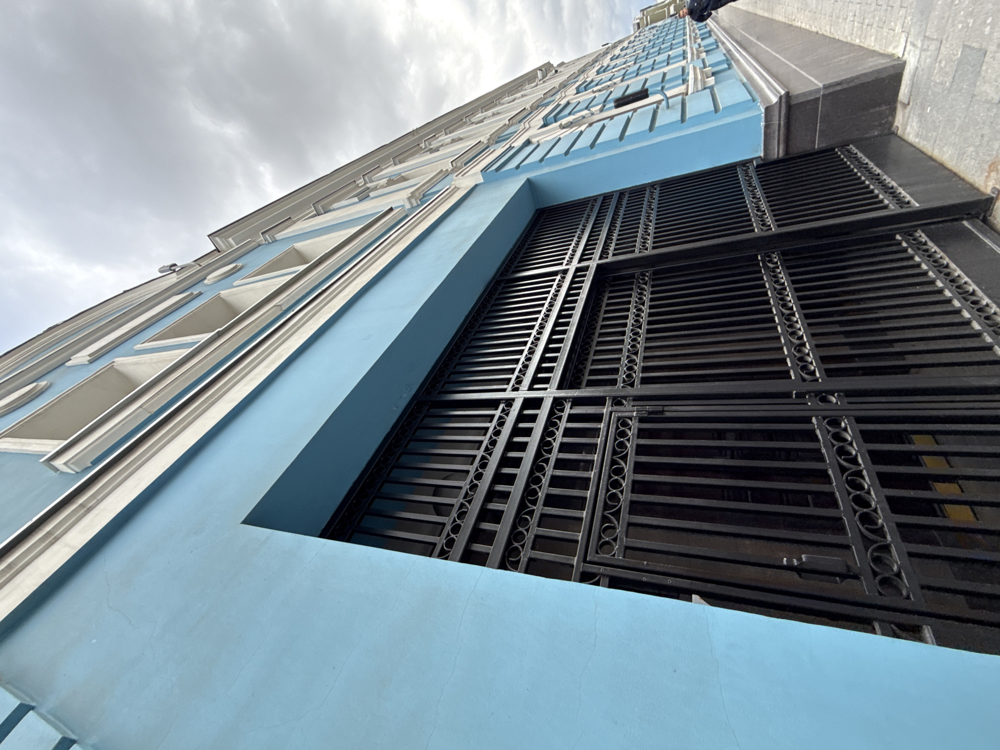 An upward, wide-angle shot captures the side of a building painted in bright blue with white trim and detailing. The building extends diagonally into the upper right corner of the frame. In the foreground, a large, black wrought-iron gate dominates the left side of the image. The gate features vertical bars and decorative horizontal elements with circular patterns. The gate appears to be partially open, revealing a dark interior. To the right of the gate, a sidewalk paved with grey and beige square stones runs along the building. The building itself has multiple windows, suggested by recesses in the blue facade. The sky above is overcast with grey clouds. A single person in a dark jacket is visible on the sidewalk in the far right of the frame, walking away from the camera. The overall scene suggests an architectural perspective of a building entrance in an urban environment. This building is located in Moscow, Russia.