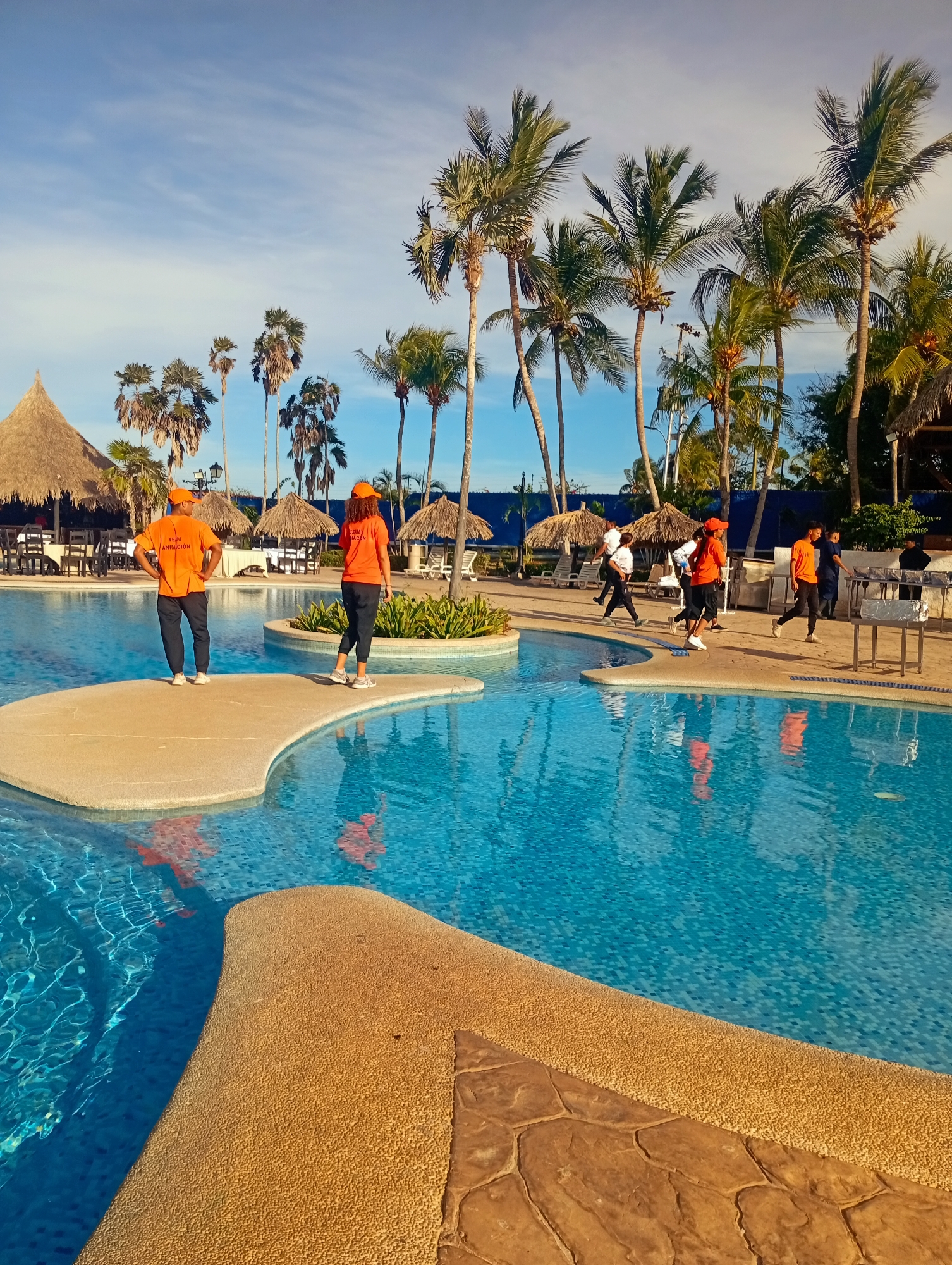The image shows a swimming pool at a resort in Playa El Agua, Venezuela. The pool is surrounded by palm trees and thatched-roof structures. Several people are walking near the pool, wearing orange shirts with "TEAM ANIMACION" printed on the back. Two people stand on a small island in the pool.