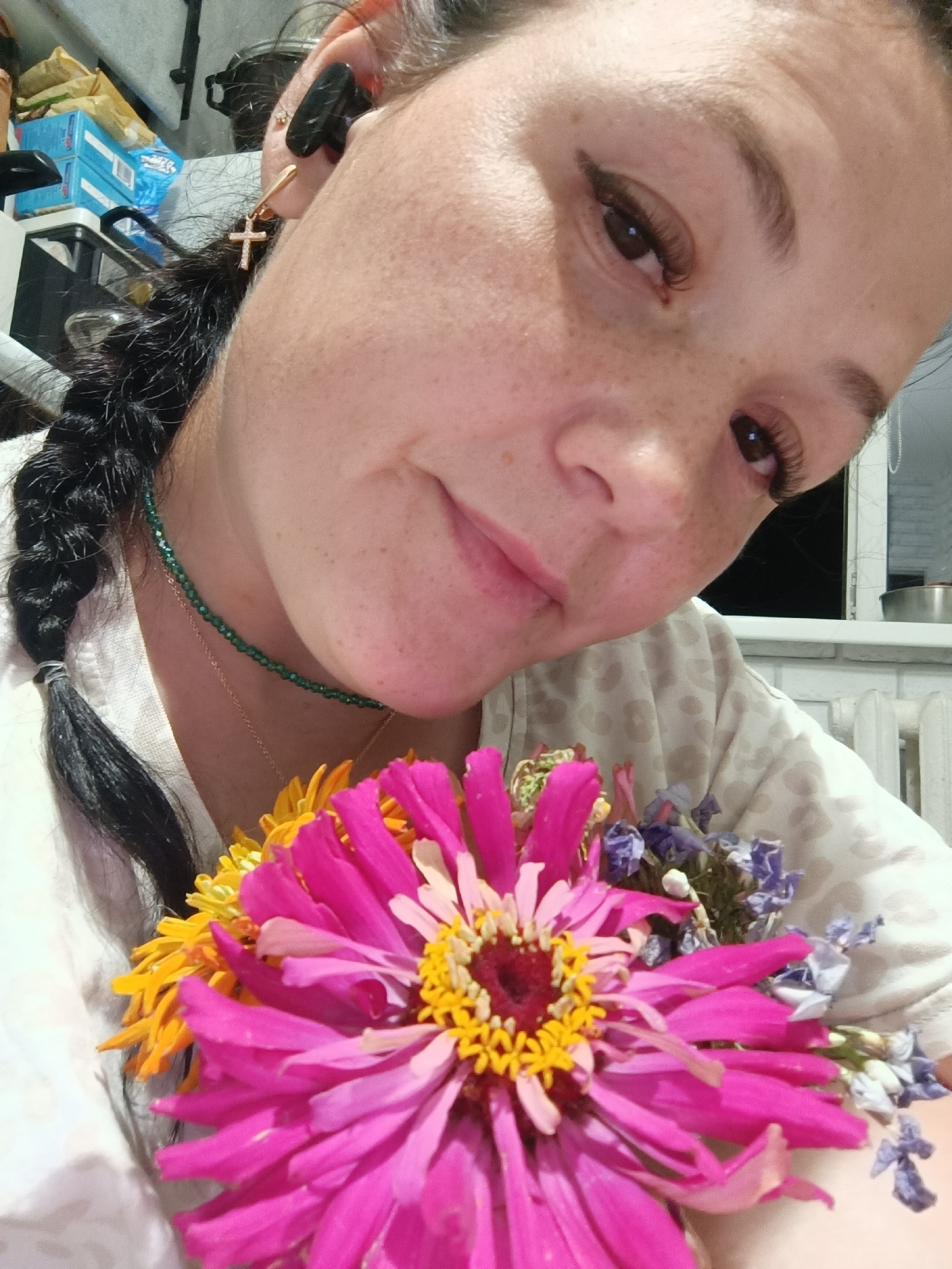 A close-up, selfie-style photograph depicts a woman with dark hair braided to her left, wearing a white shirt with a light-colored, patterned print. She has fair skin with freckles, dark eyebrows, and visible eyelashes, and appears to be wearing makeup. A cross-shaped earring with clear stones dangles from her left ear, accompanied by a black earbud. She is adorned with a thin, green beaded necklace and a delicate gold chain. The woman is holding a bouquet of flowers, predominantly a large pink zinnia with a yellow center, alongside yellow and purple dried flowers, close to her chest and face. Her head is tilted, and she is looking towards the camera with a subtle smile.
The background suggests an indoor setting, possibly a residential room in Chaikovsky, Russia. To the left, stacks of boxes are visible, one of which is blue and labeled. Further back, a dark object, possibly a shelf or cabinet, is present. To the right, a white radiator and a brick wall are visible. The lighting appears to be artificial, casting a soft glow on the woman's face and the flowers. The overall impression is that of a casual, personal moment captured indoors.