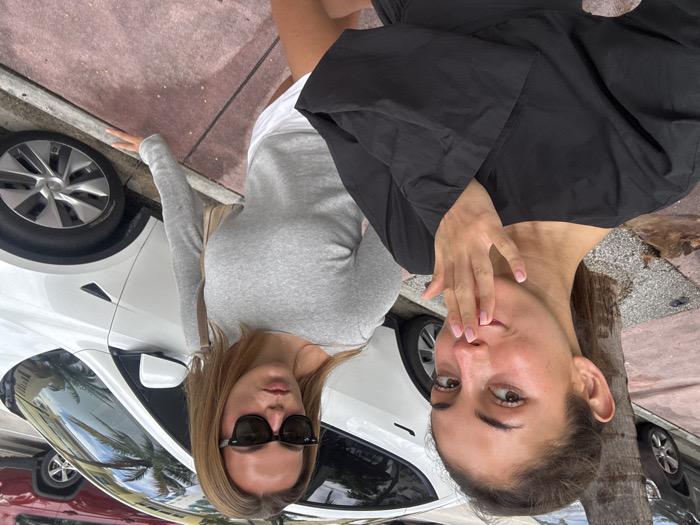 Two adult females are positioned on a reddish-pink concrete sidewalk in Miami Beach, United States, under natural daylight. The female in the foreground on the left, with light brown hair pulled back, wears a black long-sleeved top. Her right hand, featuring light pink manicured fingernails, is raised with fingers touching her lips as she looks directly at the camera. The female in the background on the right, with light brown hair down, wears dark sunglasses, a light grey long-sleeved top, and white shorts. She carries a light-colored shoulder bag and has slightly pursed lips, directing her gaze towards the camera. Behind the subjects, a white Tesla Model Y is parked parallel to the street curb, partially obscuring a dark red SUV. A tree trunk is visible on the left between the women and the parked vehicles.