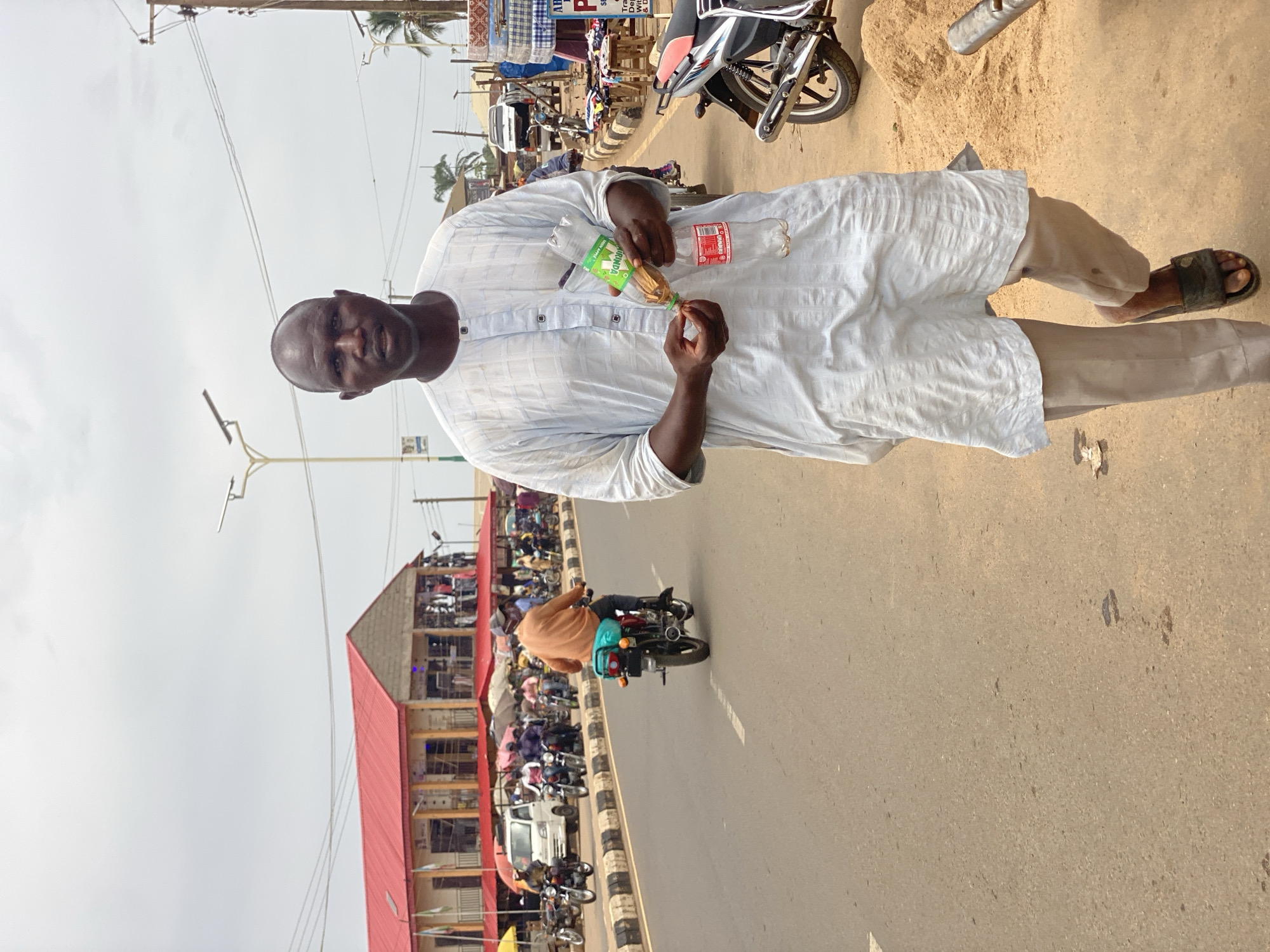 This image captures a vibrant street scene in Kafanchan B, Nigeria, during the daytime under clear, bright skies. The primary subject is a dark-skinned man standing centrally, wearing a light-colored, possibly white or off-white, patterned long tunic over light trousers and dark sandals. He is bald and looks directly at the viewer with a neutral expression. In his hands, he holds two plastic bottles: a green-labeled "AMUNDA" bottle containing a light-brown liquid, and an empty red-labeled bottle, likely a recycled Coca-Cola bottle. He appears to be in the process of pouring the liquid from the green bottle into the red one, suggesting a street vending activity such as selling oil or fuel.
The background reveals a bustling urban or semi-urban environment. An asphalt road, with a faint white line, occupies the mid-ground, populated by several people and numerous motorcycles parked or being ridden. Low-rise commercial buildings with distinct red-tiled or red-painted roofs line the street, indicating shops or establishments. Overhead utility poles and wires are visible against the bright sky, and palm trees dot the distant background. To the man's right, a large pile of sand sits on the roadside, possibly for construction or sale. The overall atmosphere suggests a busy daily life and commerce typical of a West African town. A partially obscured red sign in the background shows letters "AB" and "P".