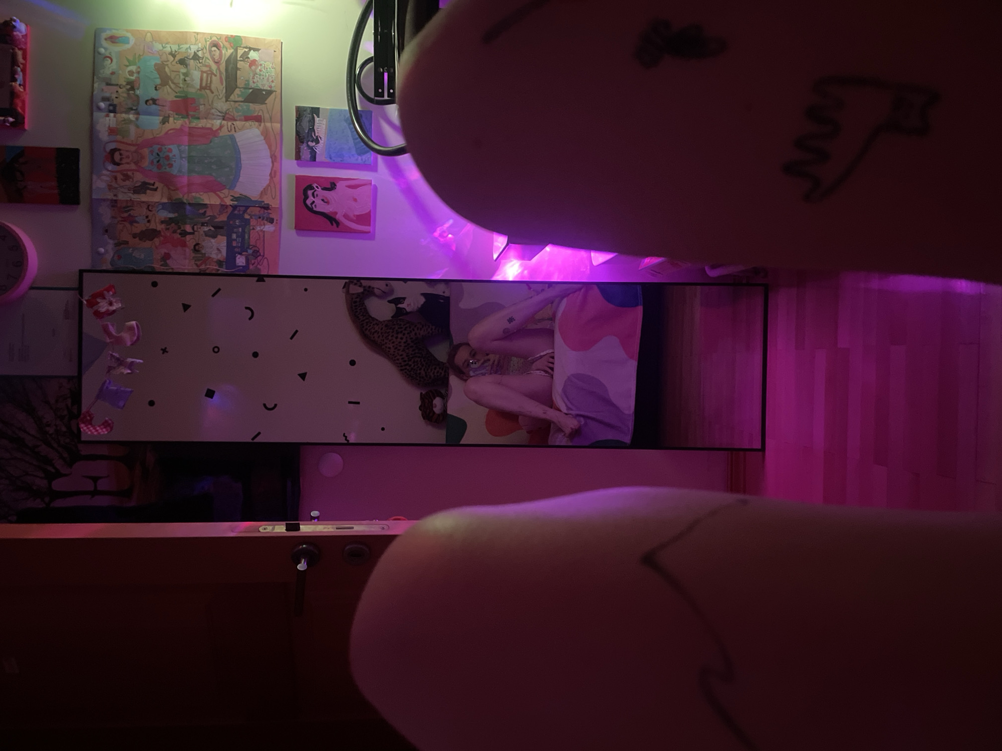 The image captures a first-person perspective, with the viewer's fingers partially obscuring the top and bottom of the frame. The top finger displays a small, stylized tattoo, while the bottom finger has a faint line tattoo.
The central focus is a wide horizontal screen, likely a television or monitor, displaying an indoor scene. On the screen, a person is seen resting or sleeping on a bed with colorful bedding featuring pink, purple, and white abstract shapes. A leopard-print object, possibly a stuffed animal or blanket, is beside them. The background of the screen scene features a light wall adorned with abstract geometric shapes like circles, crescents, and triangles.
To the left of the screen, on a wall bathed in purple ambient light, several art pieces are visible. A large poster, possibly depicting a figure reminiscent of Frida Kahlo in a vibrant dress surrounded by other scenes, is prominent. Below it, a smaller rectangular artwork with red and blue tones hangs. Above and to the right of the large poster, two smaller square paintings are mounted; one features blue hues, and the other, a pink background with a stylized figure. A pink circular clock is partially visible on the far left.
The room is illuminated by prominent purple LED strip lighting, particularly casting a strong glow on the right side of the wall, suggesting an evening or nighttime setting. A faint green light source is also visible in the upper left corner. Below the screen, a wooden desk or table surface with metallic accents is present. The overall setting appears to be a personal space, possibly a bedroom, characterized by artistic decoration and mood lighting. No discernible text is visible in the image. The location context for this scene is Belgrade, Serbia.