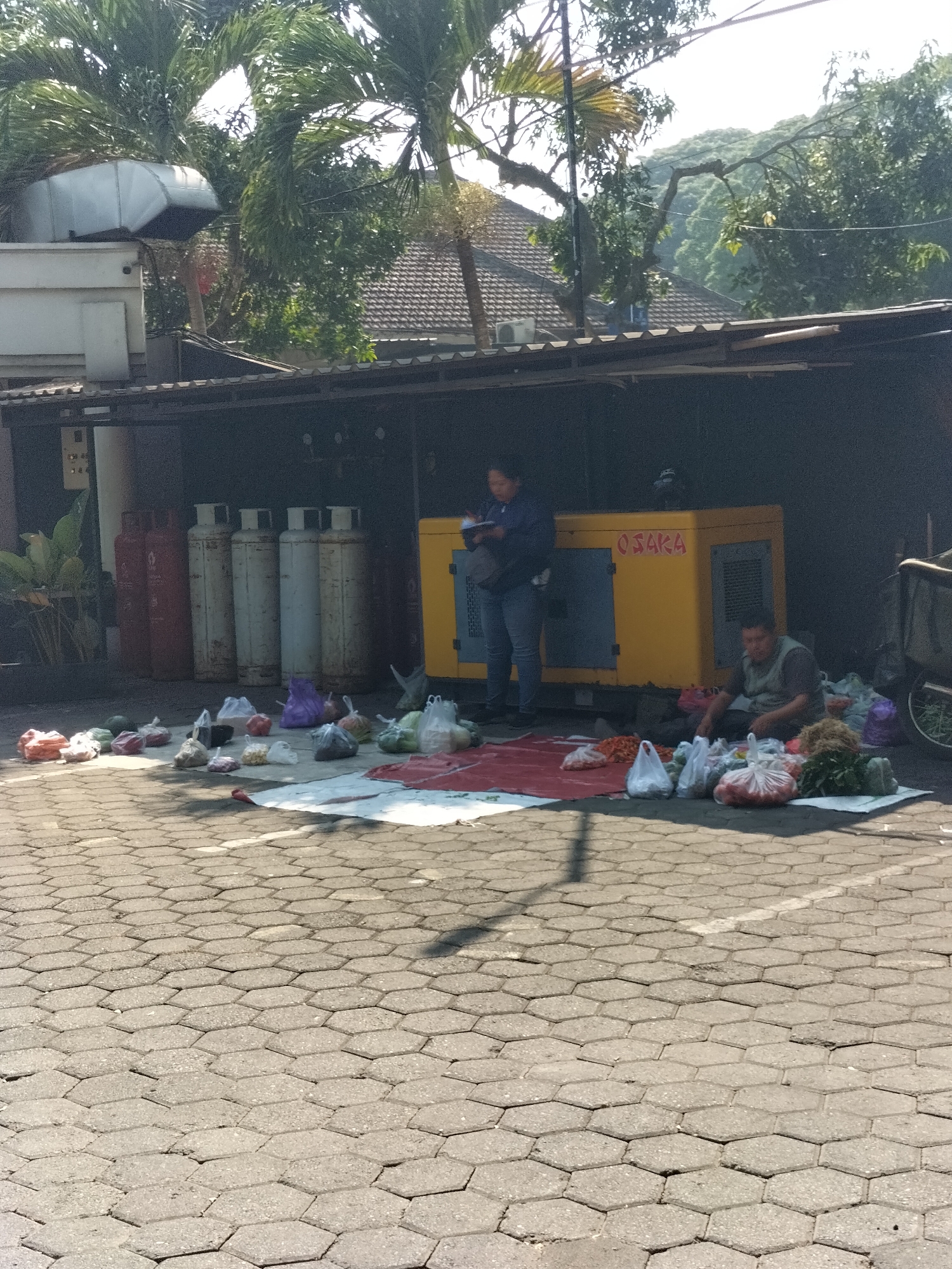 An outdoor scene depicts a market stall set up in front of a large yellow generator labeled "OSAKA." The ground is paved with hexagonal interlocking stones. Several people are present. In the background, a woman in a blue shirt and jeans stands near the generator, looking at a device in her hand. A man in a dark green vest and pants sits on the ground to the right of the generator, surrounded by various items. In the foreground and spread out on the ground are numerous plastic bags filled with what appear to be produce and other goods, suggesting a temporary market or roadside sale. Behind the generator, several large gas cylinders are visible, some red and some white. Tall palm trees and dense foliage are visible in the background, indicating a tropical or subtropical environment. The overall setting suggests a commercial area, possibly in a city like Malang, Indonesia, where such informal markets are common.