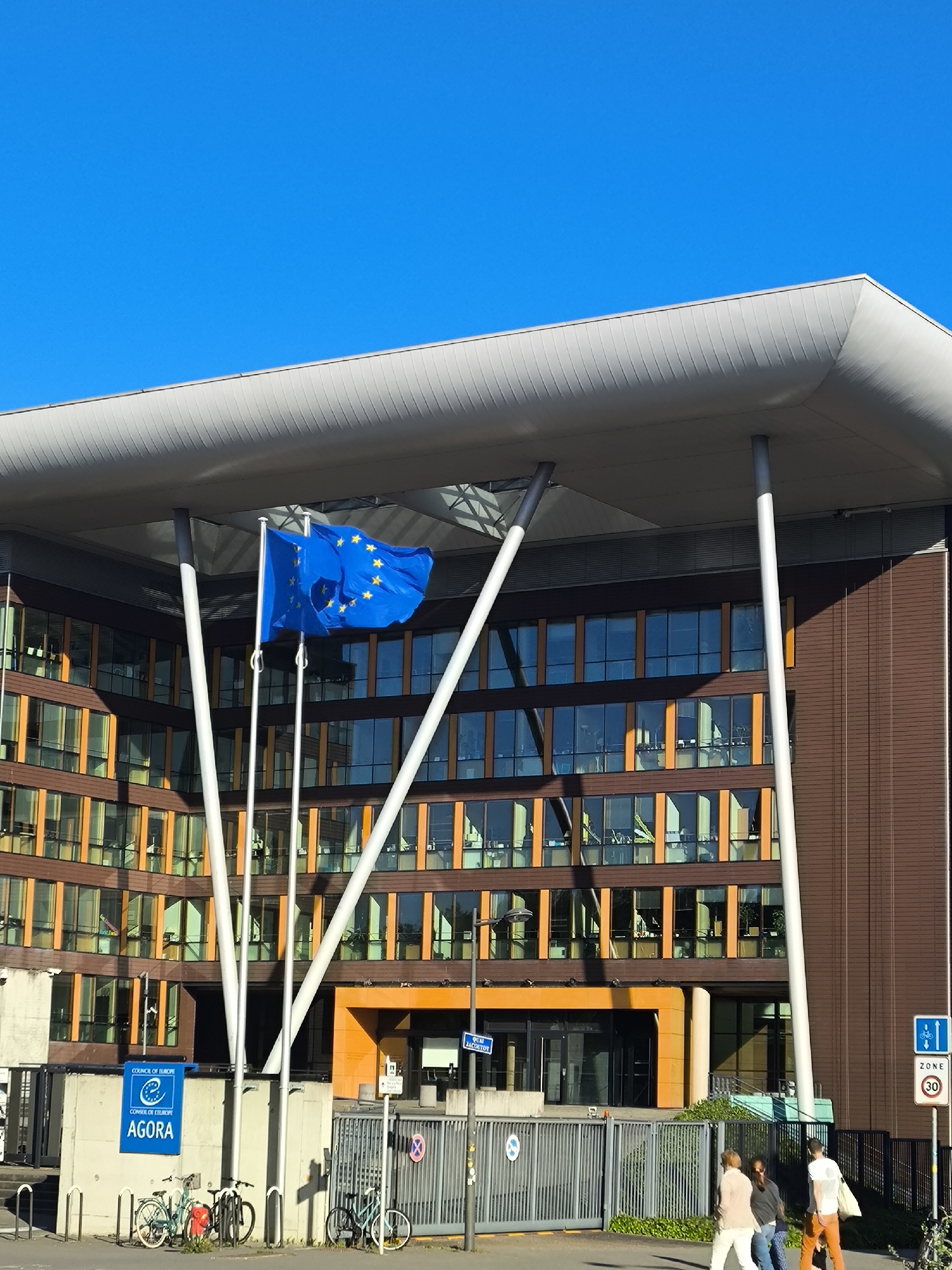 The image shows the Council of Europe's Agora building in Strasbourg, France, under a clear blue sky. The building is a modern structure with a gray roof supported by white angled pillars. The facade features horizontal bands of brown panels and numerous windows with orange frames. The European Union flag flies in front of the building.
A gray metal fence runs along the base of the building, with several bicycles parked near a sign that reads "COUNCIL OF EUROPE" and "CONSEIL DE L'EUROPE AGORA". Several people are walking on the sidewalk in front of the fence, with one wearing an orange pants. A blue sign with speed limits is mounted on a pole next to the fence.