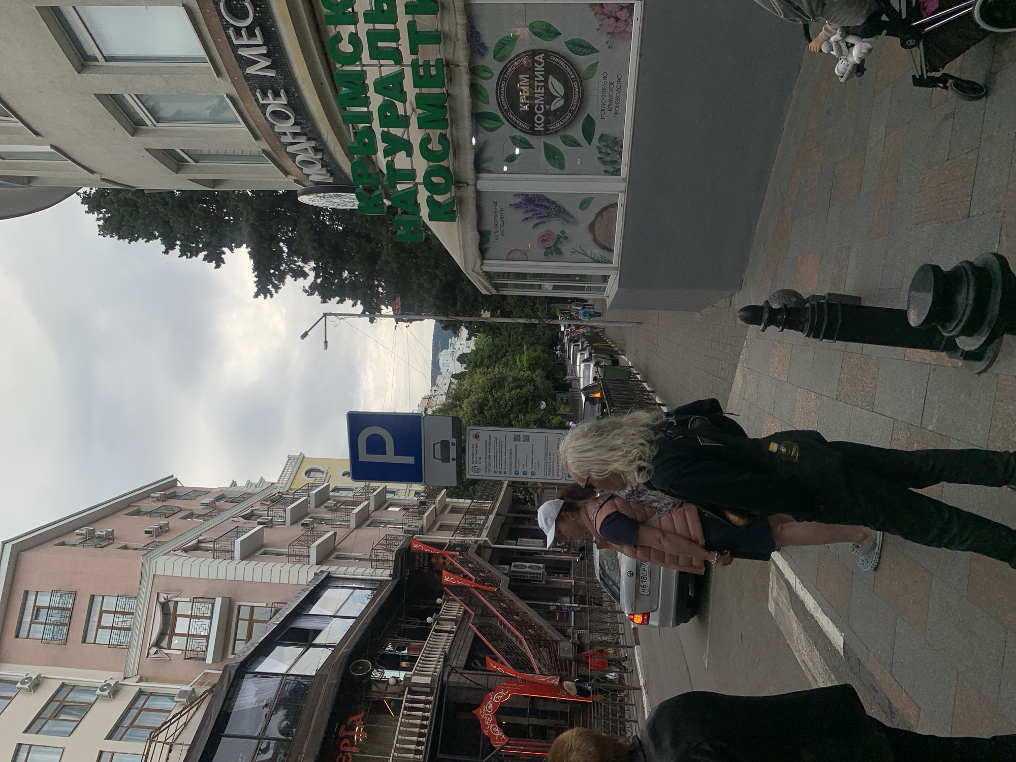 The image, when properly oriented, depicts an urban street scene on an overcast day, featuring buildings, pedestrians, and parked cars. In the foreground, two individuals are walking on a paved sidewalk. One person, with long, wavy blonde hair, is dressed in a black jacket and black pants, with a small yellow item visible in their back pocket, and light blue Crocs on their feet. Adjacent to them is another person wearing a white baseball cap, a pink vest over a dark top, and dark pants. A silver sedan with the license plate H618CP 75 is parked nearby.
On the left side of the street, several multi-story buildings line the sidewalk. A prominent building features a large sign with green Russian text, reading "КРЫМСКАЯ НАТУРАЛЬНАЯ КОСМЕТИКА" (Crimean Natural Cosmetics). Its display window is adorned with a circular logo stating "КРЫМ КОСМЕТИКА" (Crimea Cosmetics) and phrases such as "100% НАТУРАЛЬНЫЕ ИНГРЕДИЕНТЫ" (100% Natural Ingredients), "УНИКАЛЬНО НАТУРАЛЬНО КАЧЕСТВЕННО" (Unique Natural Quality), and "ИСКЛЮЧИТЕЛЬНО КРЫМСКОЕ ПРОИЗВОДСТВО" (Exclusive Crimean Production), surrounded by illustrations of plants and leaves. Part of another sign on an adjacent building reads "ОДНОЕ МЕС".
Further down the street, more buildings with varied architectural styles are visible, some displaying red decorative elements. A blue parking sign with a white 'P' symbol and another informational sign with QR codes and Russian text are present. The street is lined with parked cars and mature trees. The sky is predominantly cloudy, and the overall lighting suggests daytime. Black bollards and a lamp post are visible along the sidewalk. A partial view of a baby stroller is seen on the far right.