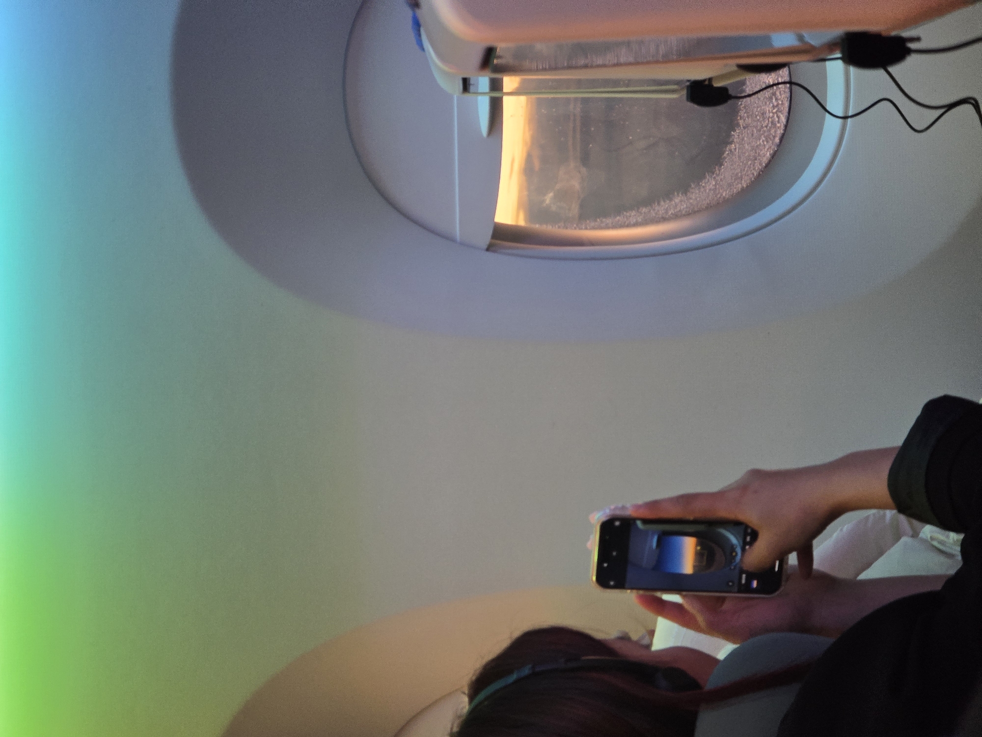 The image captures a first-person perspective from within an airplane cabin, focusing on a person taking a photo of the window view with a smartphone. The scene is dominated by the light-colored interior of the aircraft.
In the foreground, at the bottom right, the back of a person's head, shoulder, and hands are visible. The person has dark hair and appears to be wearing headphones or a headset. They are holding a silver or light-colored smartphone horizontally, pointing it towards the airplane window. The screen of the smartphone clearly displays a reflection of the view outside the window.
The central subject is an oval-shaped airplane window, which reveals a vibrant outdoor scene. Frost or ice crystals are visible along the edges and lower portion of the window pane, suggesting a high altitude and cold conditions. Through the window, warm, bright orange and yellow light, indicative of either sunrise or sunset, illuminates the exterior. This light reflects strongly on parts of the window and the ice. Some dark wires or cables are visible hanging near the upper right corner of the window.
The interior of the plane walls is smooth and light-toned. On the left side of the image, there's a distinct light gradient, transitioning from bright cyan/blue at the far left to a more subdued green and then a neutral light gray towards the center. This gradient might be due to interior lighting or reflections. The overall impression is one of capturing a beautiful moment during flight. There is no visible text in the image.