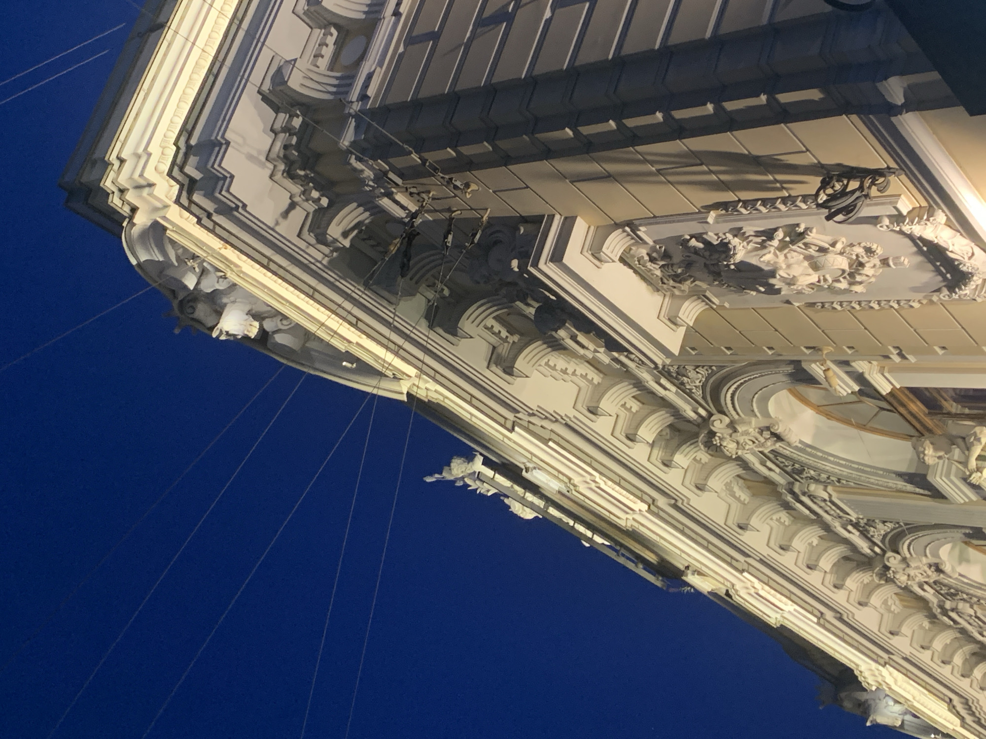 The image shows an ornate building facade in Saint Petersburg, Russia, under a dark blue night sky. The building features intricate architectural details, including decorative moldings, carvings, and what appear to be sculptural elements, illuminated by warm lighting. Several thin wires stretch across the sky, likely utility or electrical lines, adding a utilitarian contrast to the elaborate architecture. The perspective is from a low angle, looking up at the upper floors and roofline of the building. No people are visible in the image.