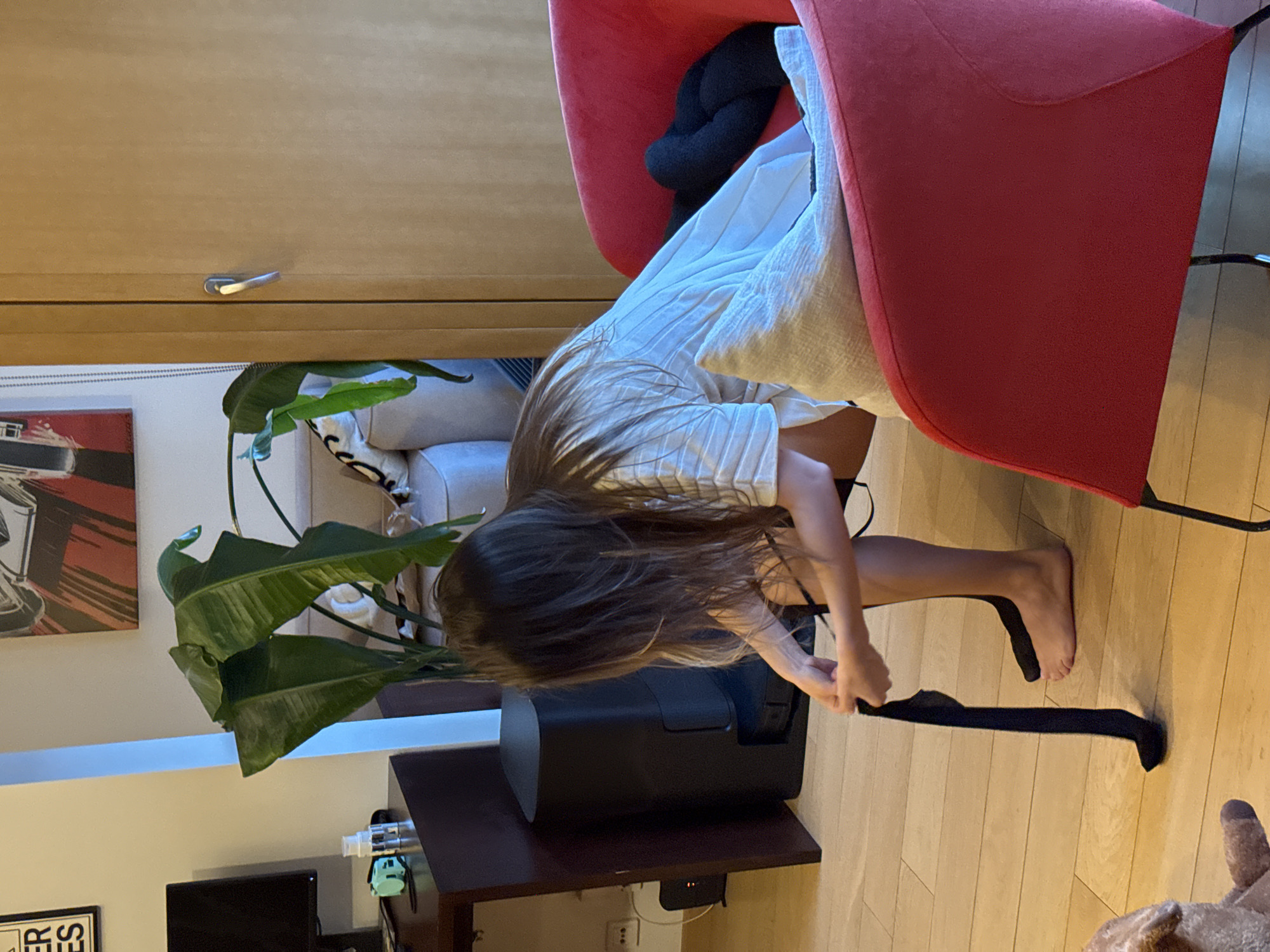 A young girl with long brown hair is pulling on a black stocking while sitting in a red chair. The scene is indoors, in a room with wood floors. In the background, there is a cabinet, a plant, and part of a couch. The presence of an electrical outlet of a specific type suggests this is likely in Europe, potentially Warsaw, Poland.