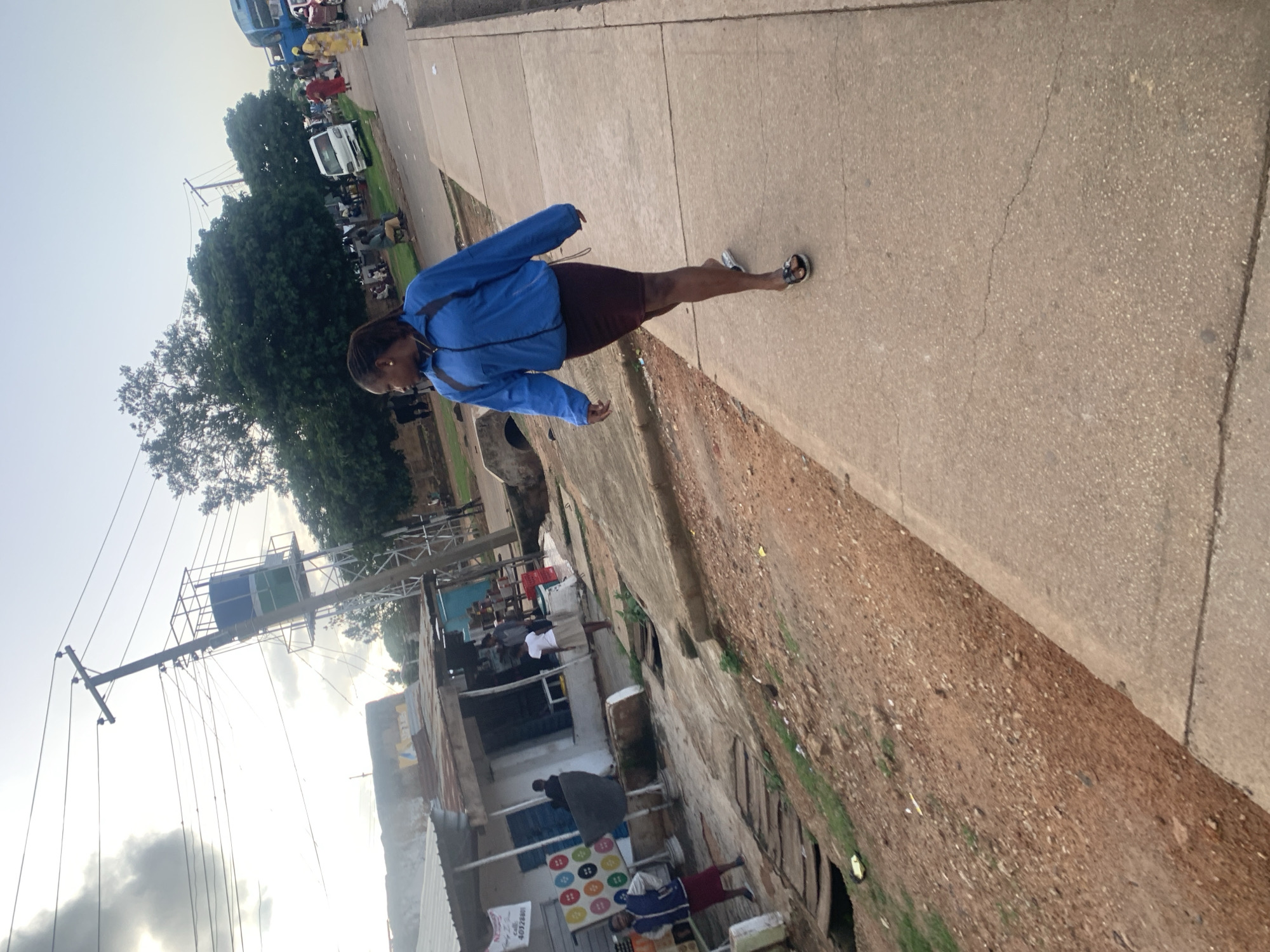 The image depicts a street scene in Jos, Nigeria. In the foreground, a woman wearing a blue jacket and a dark skirt walks on a paved sidewalk. She is looking to her left. Behind her, to the left, is a roadside area with dirt, some vegetation, and what appears to be a drainage system. Further back, a row of small shops or market stalls with corrugated metal roofs are visible. People are seen in and around these stalls, some standing and some sitting. A utility pole with electrical wires and a water tank structure stands prominently in the mid-ground. In the background, trees and more distant buildings can be seen. Further down the street, there are vehicles, including a bus and vans, and more people. The sky is overcast. The overall impression is of a bustling urban or semi-urban environment.