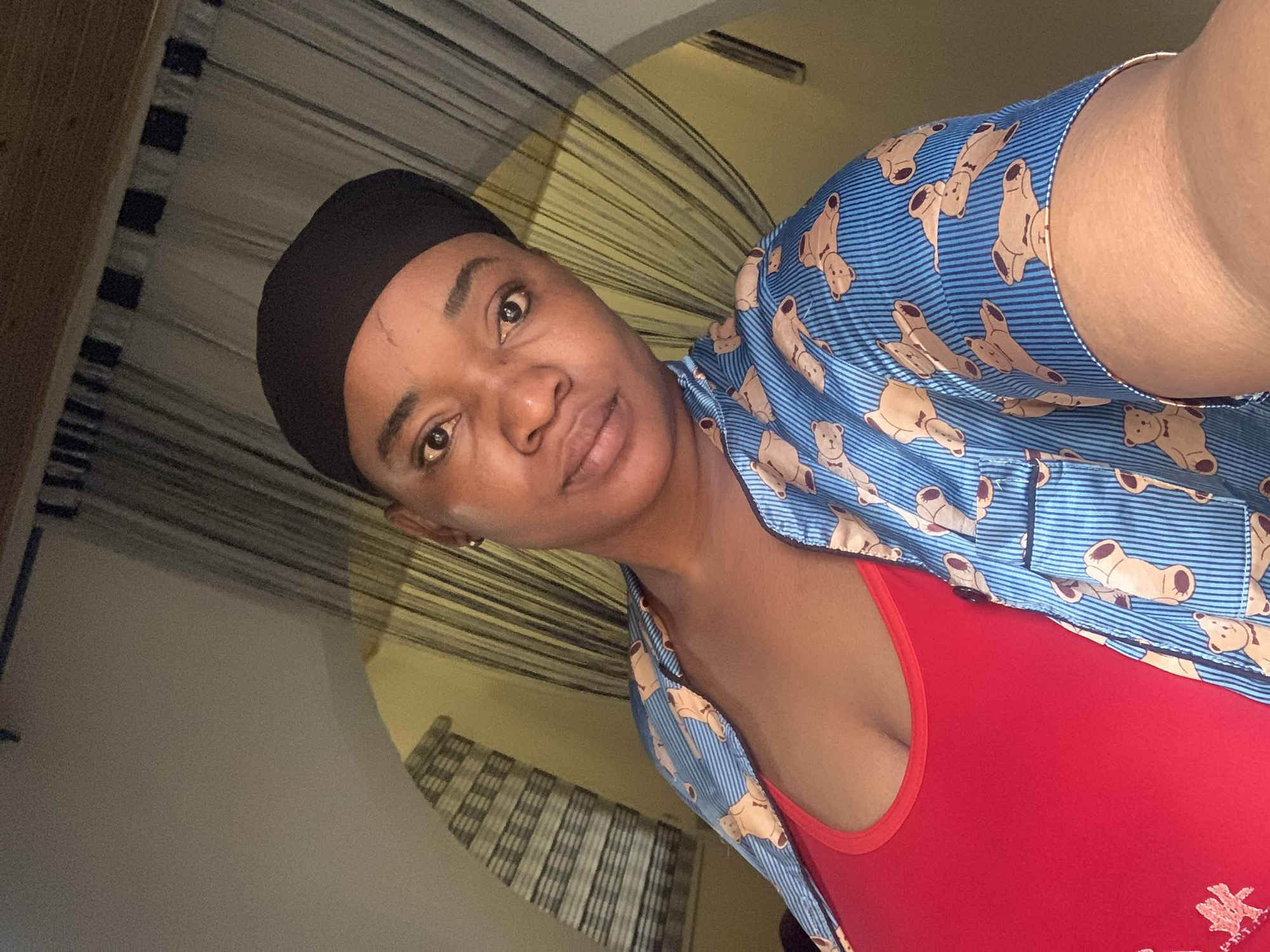 A woman with dark hair covered by a black cap is facing the camera. She is wearing a red tank top under a light blue button-down shirt with a teddy bear pattern. The background shows striped curtains and a light-colored wall. The image does not provide verifiable data about the city of Barakin Akawo, Nigeria.