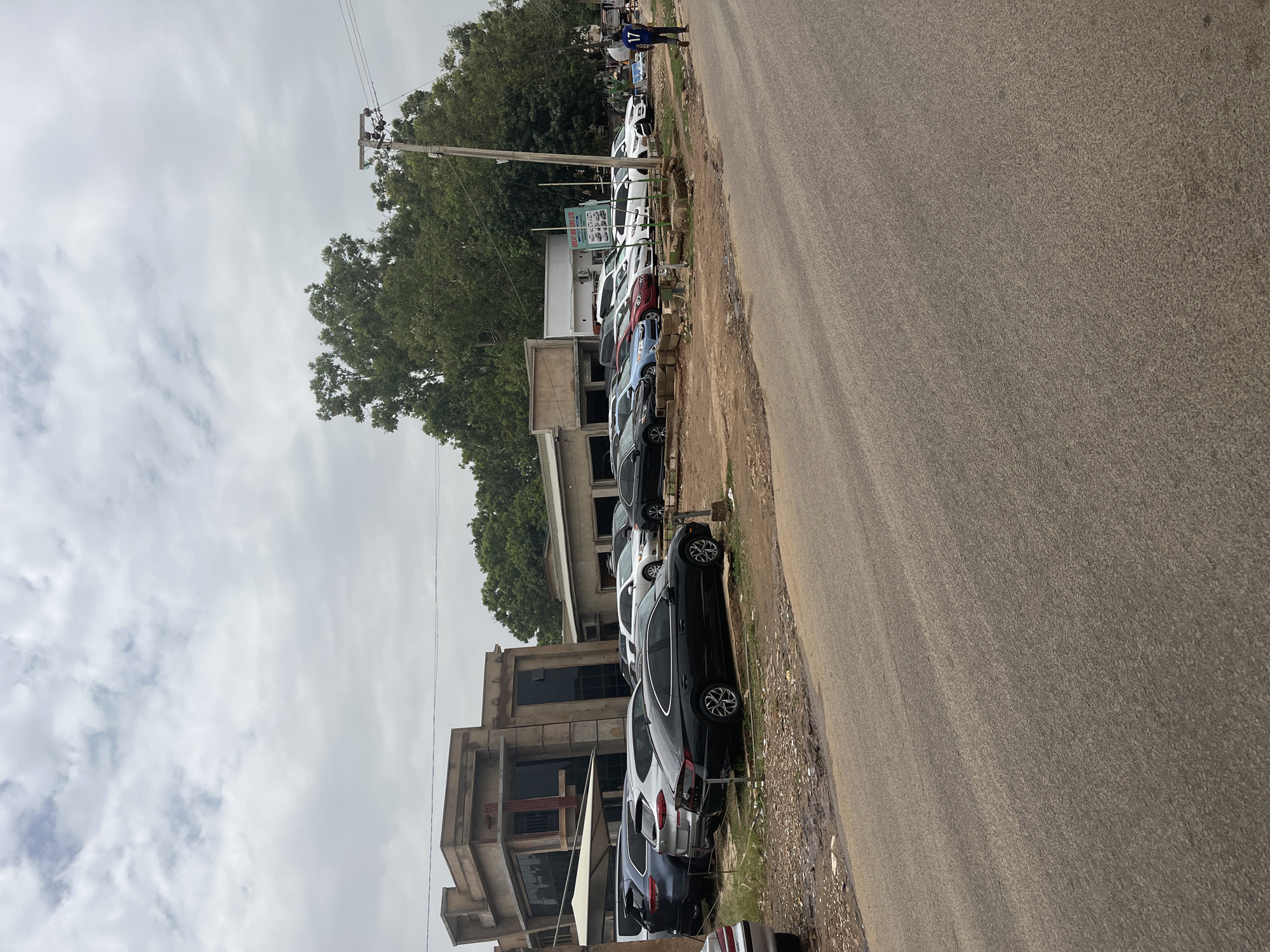 The image shows a street in Jos, Nigeria. A row of parked cars lines the side of the road, adjacent to a multi-story building. The building appears to have a modern architectural design. A sign labeled "BEST GRADES LTD" is visible. A person is walking on the roadside further in the distance. Utility poles and power lines are seen overhead, against a cloudy sky. The road surface is asphalt and appears to be in reasonably good condition.