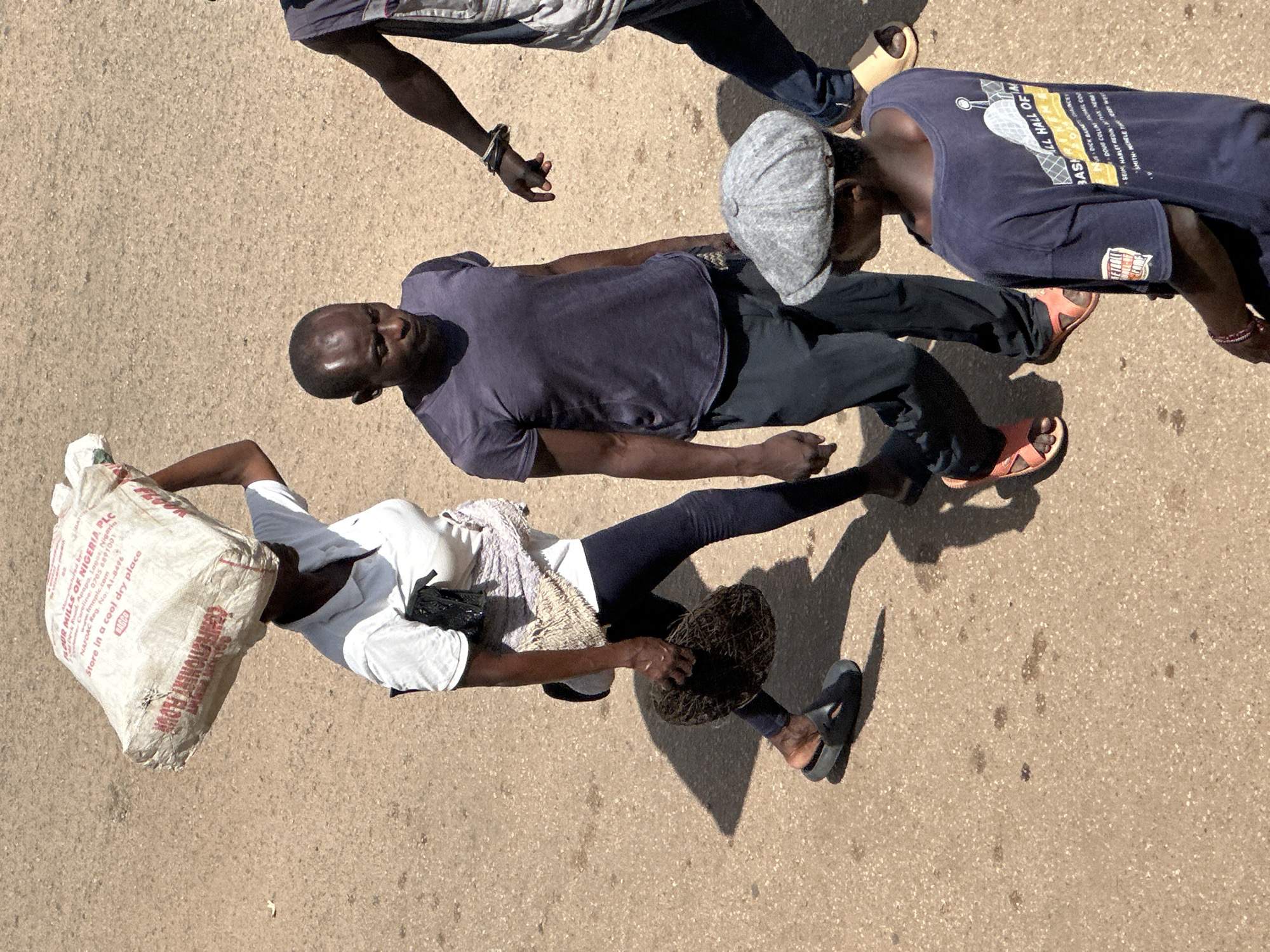 In Bukuru, Nigeria, four individuals are captured in an overhead shot, walking on a light brown, textured surface that appears to be asphalt or a similar paved area with scattered dark spots.
A woman in the foreground on the left is carrying a large, white sack of flour on her head. The sack is printed with "FLOUR MILLS OF NIGERIA PLC" and instructions. She wears a white polo shirt and dark leggings, with a black purse or bag slung over her shoulder. She holds a round, dark, woven object in her right hand and wears dark flip-flops.
To her right, a man with a shaved head is walking. He is wearing a dark purple t-shirt and dark pants. His gaze is directed downwards.
Further to the right, another man is visible from the back, wearing a dark sleeveless vest over a t-shirt and dark pants. He has bracelets on his left wrist.
In the lower right of the frame, a person wearing a grey cap and a dark blue t-shirt with text and graphics on the back is partially visible, facing away from the camera. This individual also appears to be walking.
The lighting suggests it is daytime, casting distinct shadows on the ground. The overall scene depicts everyday life and movement in an outdoor urban environment.