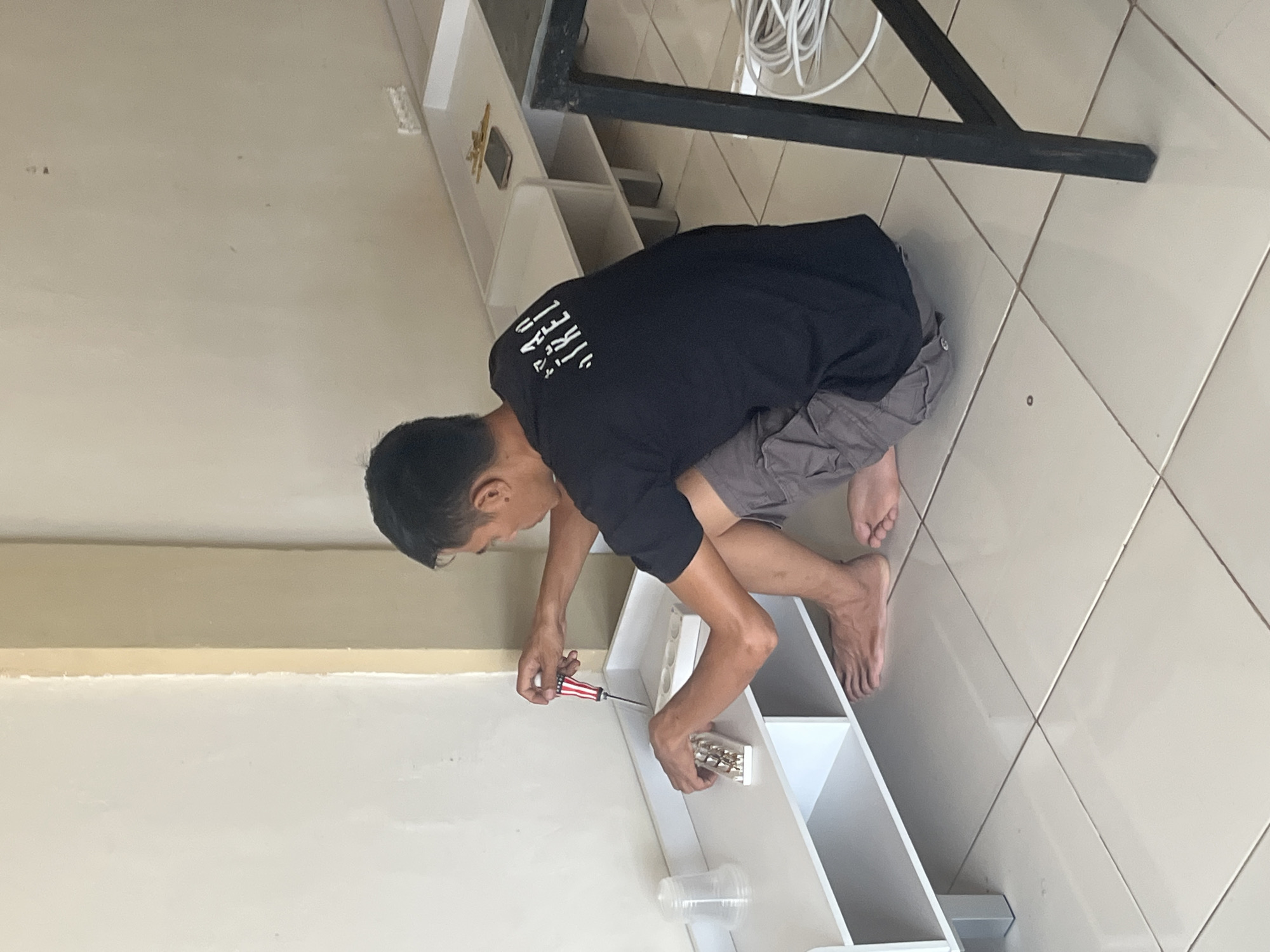 In Sekayu, Indonesia, a man is assembling a white shelving unit indoors. He is crouched on the tiled floor, wearing a black t-shirt and grey shorts. In his right hand, he holds a screwdriver with a red, white, and blue handle, and in his left, he holds a plastic case containing screws. The shelving unit is modular, with several compartments visible, and is positioned against a pale yellow wall. To the right of the shelving unit, a dark metal table leg and coiled white cables are visible, suggesting the presence of electronic equipment or an ongoing setup. The floor is made of large, light-colored tiles, and the overall lighting indicates an indoor environment, likely a room in a house or a small shop.