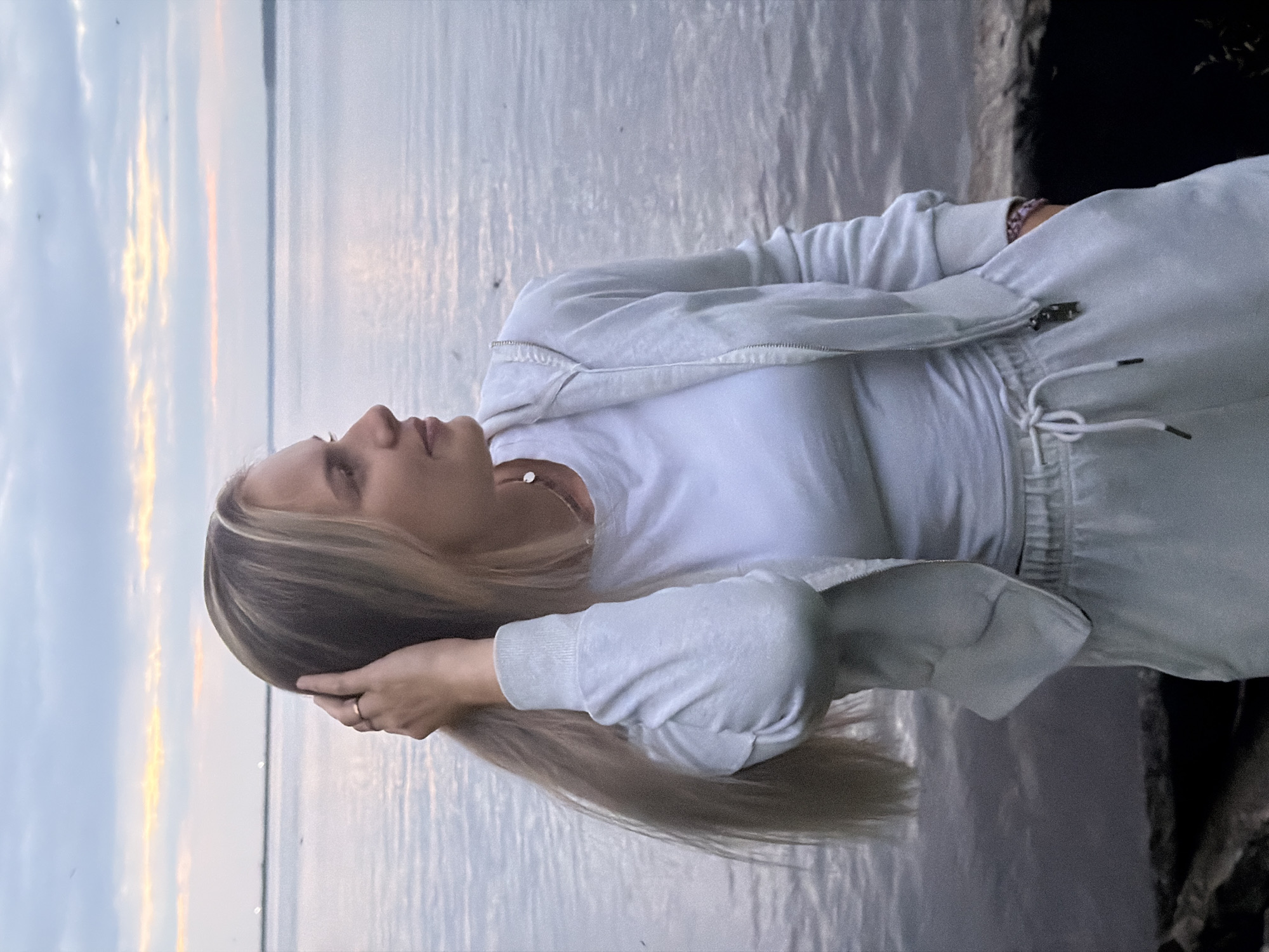 A young woman with long blonde hair is standing on the shore of a body of water. She is wearing a white t-shirt and a light blue tracksuit. Her right hand is in her hair, and she is looking to her right, away from the camera. The sky is cloudy with streaks of orange and pink, suggesting it is either sunrise or sunset. The water is calm, reflecting the sky. The foreground shows a dark, rocky shoreline. This scene appears to be located at the Kama River embankment in Chaikovsky, Russia, a city known for its industrial and recreational areas along the river.