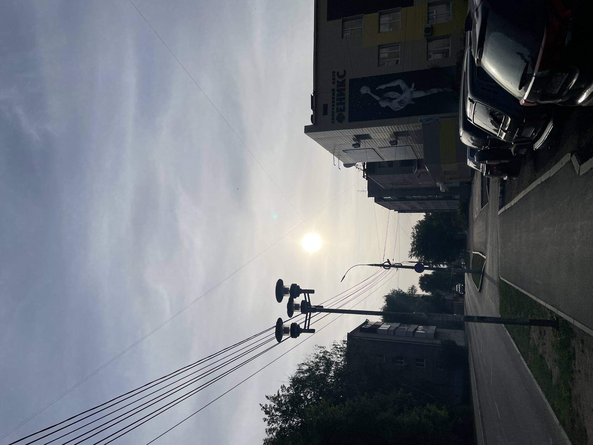 The image shows a street in Izhevsk, Russia, on a sunny day. Tall buildings line one side of the street, with cars parked along the curb. There is a mural on one building that reads "Спортивный Клуб Феникс" ("Phoenix Sports Club") and depicts a female volleyball player. Utility poles and wires run along the street. A sidewalk is visible next to the road. The sky is hazy.
