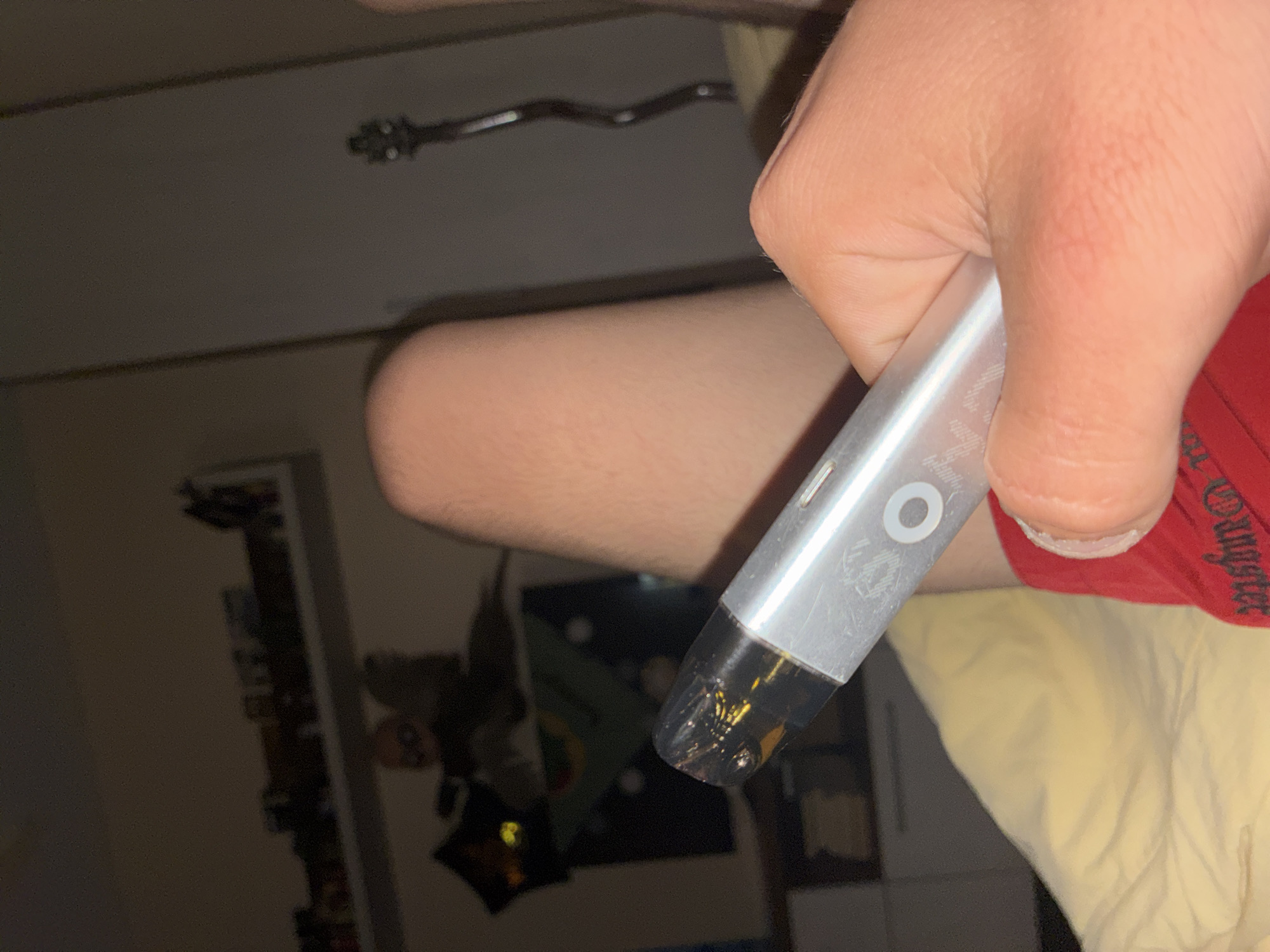 The image captures a close-up, dimly lit indoor scene featuring a person's hand holding a silver vape device. The right hand, with visible skin texture and creases, firmly grips the vape. The device itself is metallic silver, showing some minor wear or scratches, and features a prominent circular button or indicator light on its side. The mouthpiece end is transparent and dark, revealing a yellowish liquid, likely vape juice, within the pod.
In the blurred background, parts of the person's body are visible. A section of a bare leg or thigh is seen on the left. On the right, a red garment is present, displaying partially visible white text that reads "™" and "gangster," suggesting a brand name or design on clothing such as shorts or a t-shirt. Below this red fabric, a light yellow or cream-colored fabric, possibly bedding or a cushion, fills the lower right corner of the frame.
The ambient lighting is low, indicative of an indoor setting during the evening or night. Further in the soft-focus background, indistinct shapes suggest furniture, possibly a shelf or cabinet, and a glinting metallic object, which could be part of an exercise machine or a structural element, is visible. The image depicts a casual moment of a person holding a vape, set against a domestic backdrop in what is contextually understood to be Varna, Bulgaria.